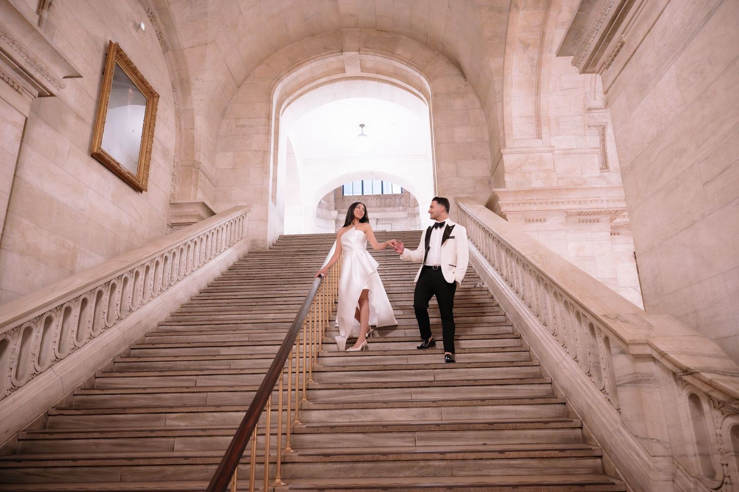 Couple runs hand in hand down elegant white marble staircase in historic building with arched window.