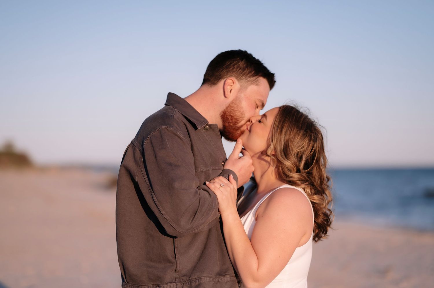 Couple sharing a romantic kiss on a sandy beach at sunset, with calm ocean waters in the background.
