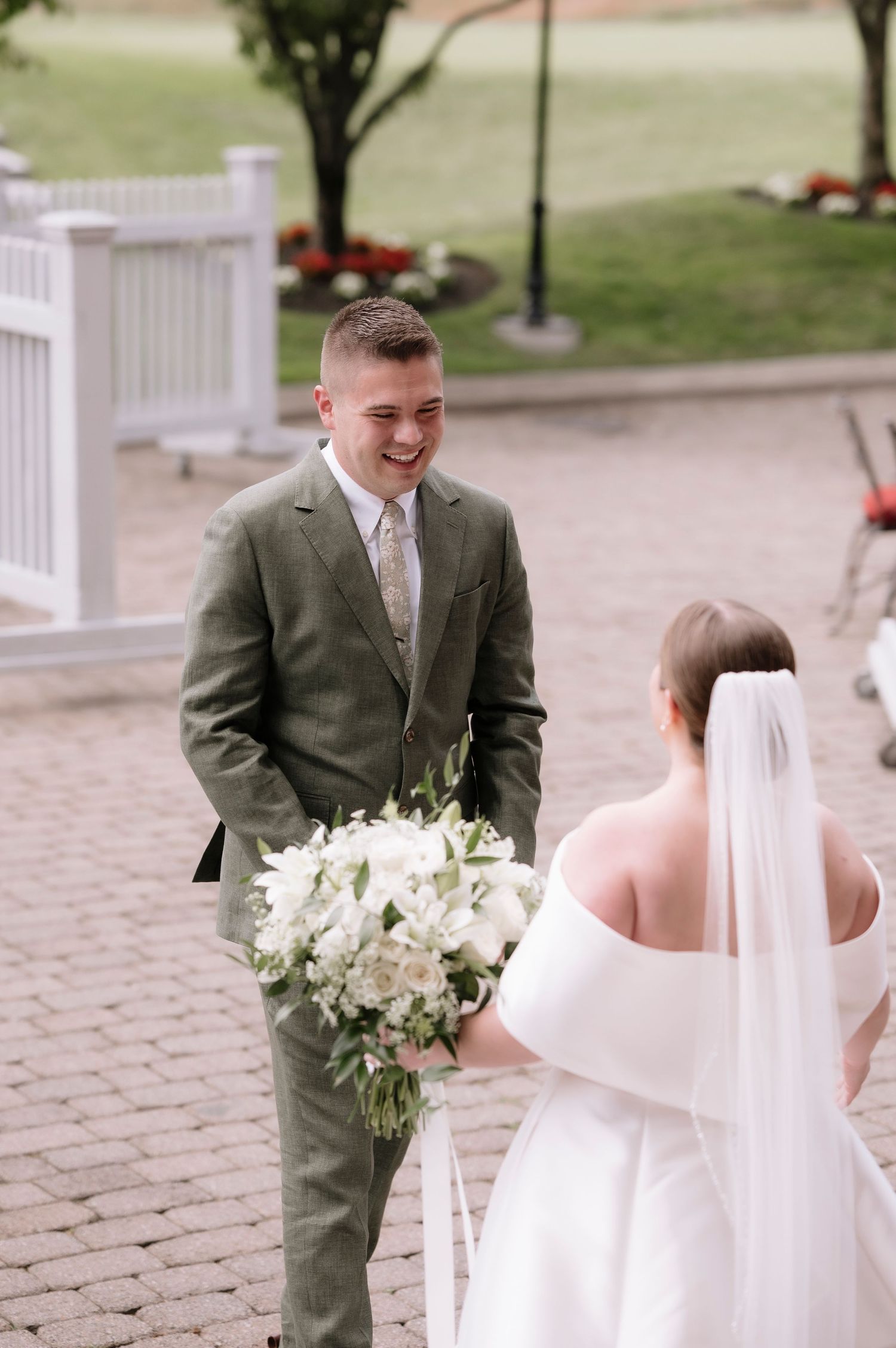 Groom in olive green suit smiling at bride in white off-shoulder gown holding white floral bouquet during first look.