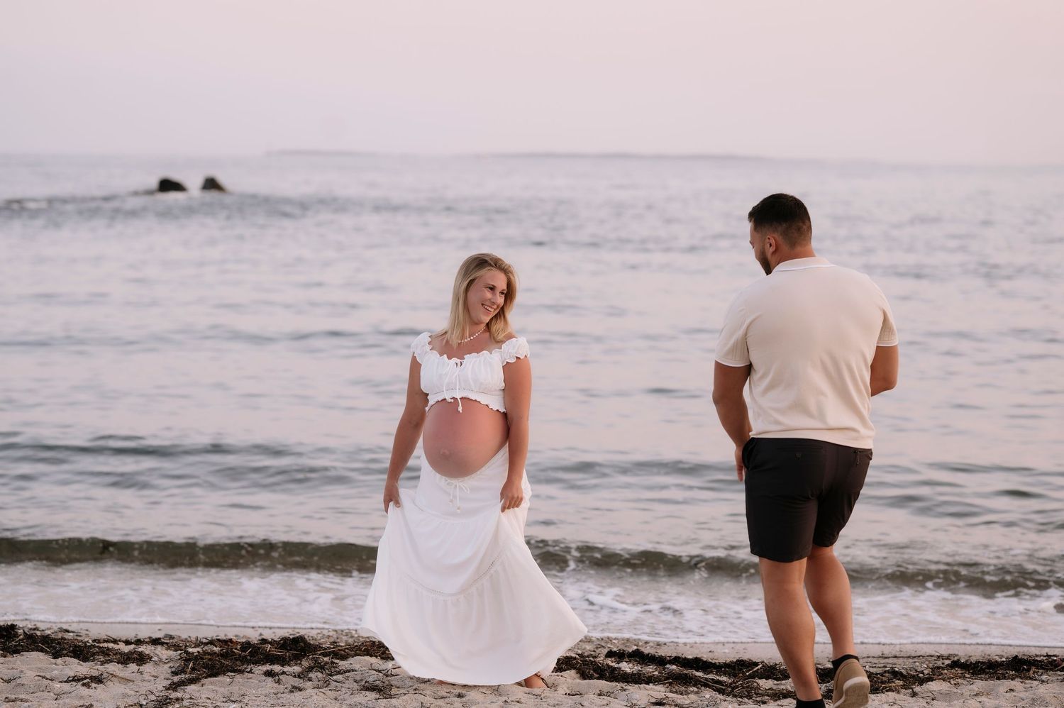 Pregnant woman in white dress stands on beach while partner walks toward her at sunset.