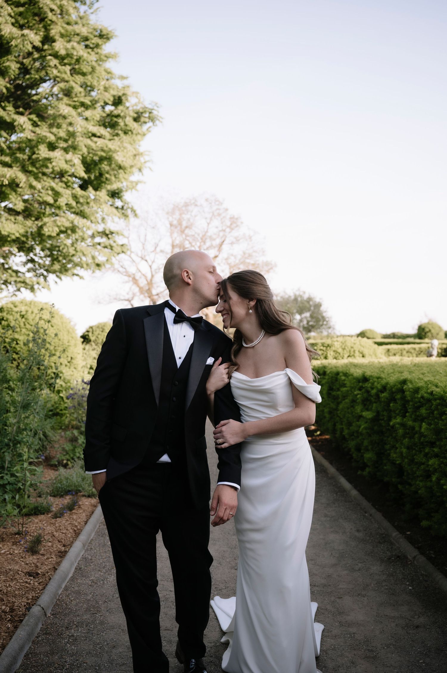 A wedding couple embraces while walking along a garden path bordered by manicured hedges on a sunny day.