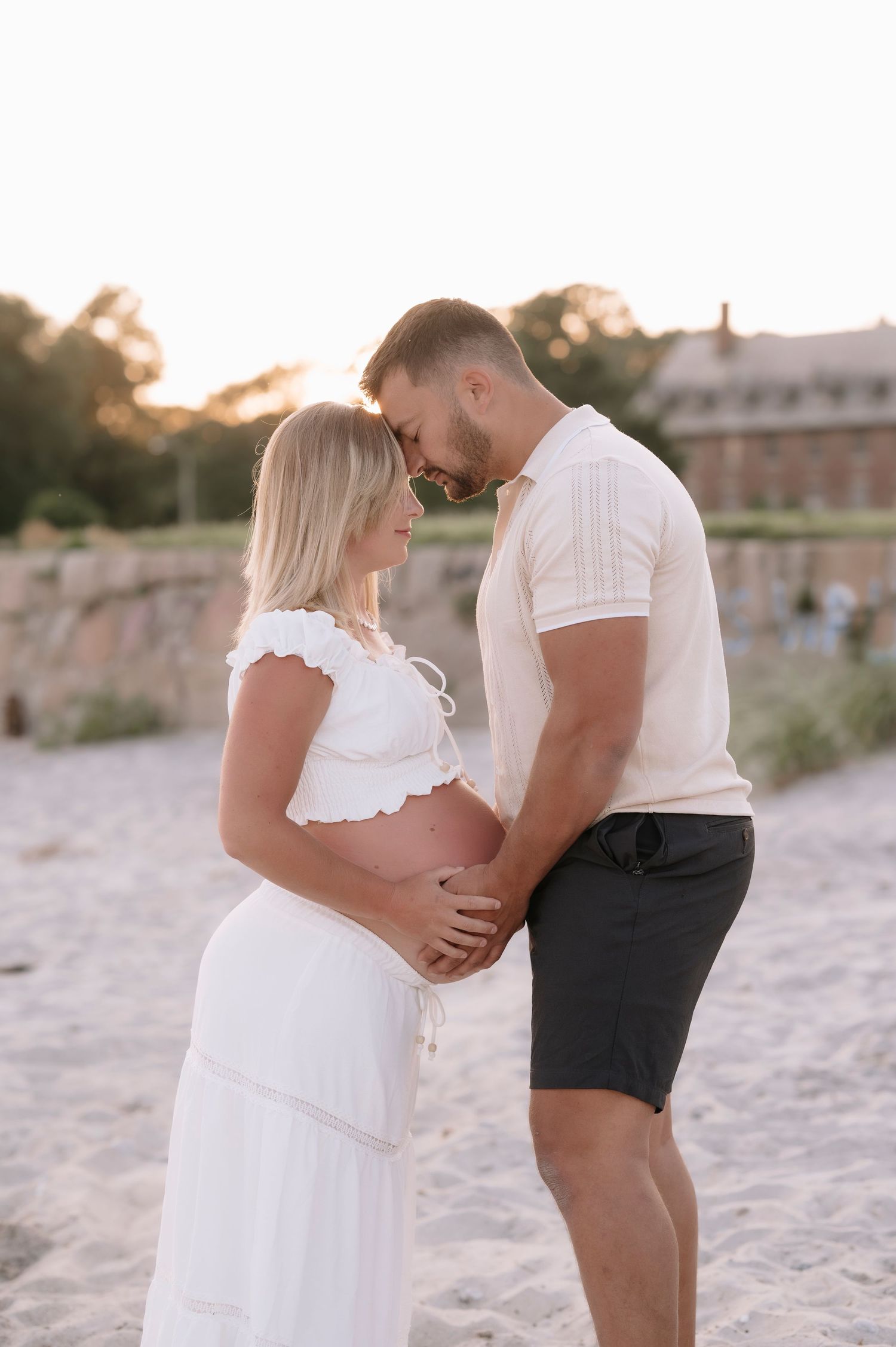 Couple sharing a tender maternity moment on a sandy beach at golden hour, man cradling pregnant womans belly.