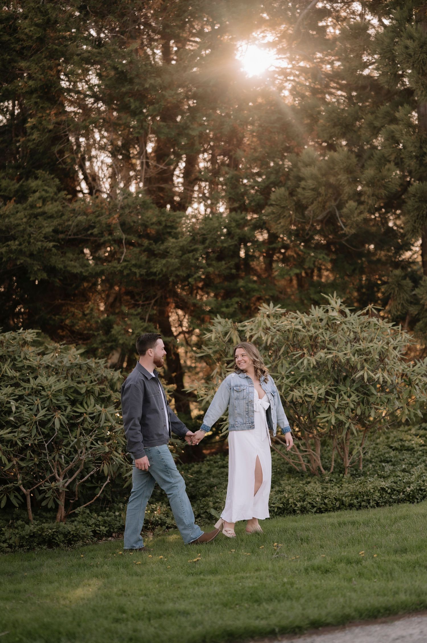 Couple holding hands walking through lush green garden at golden hour sunset, woman in white dress and denim jacket.