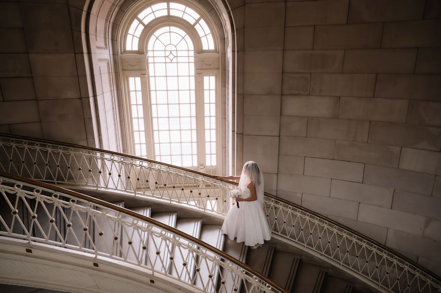 Bride in white dress standing on elegant curved staircase by arched window in grand stone building.
