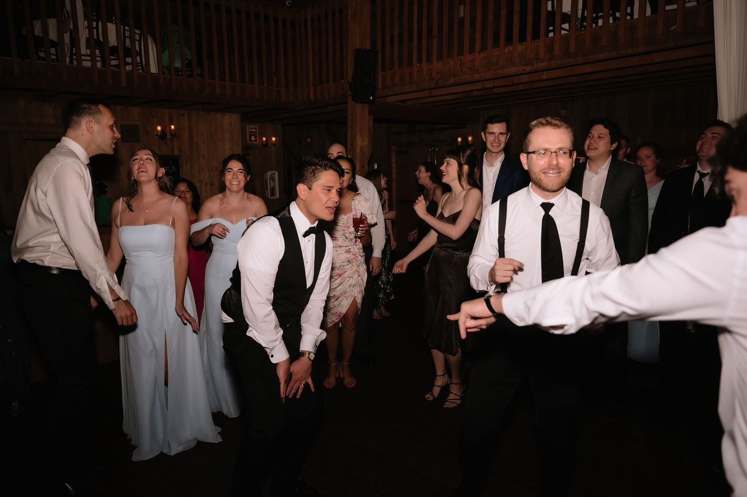 Groomsmen and wedding guests dancing energetically inside a rustic barn wedding venue reception.