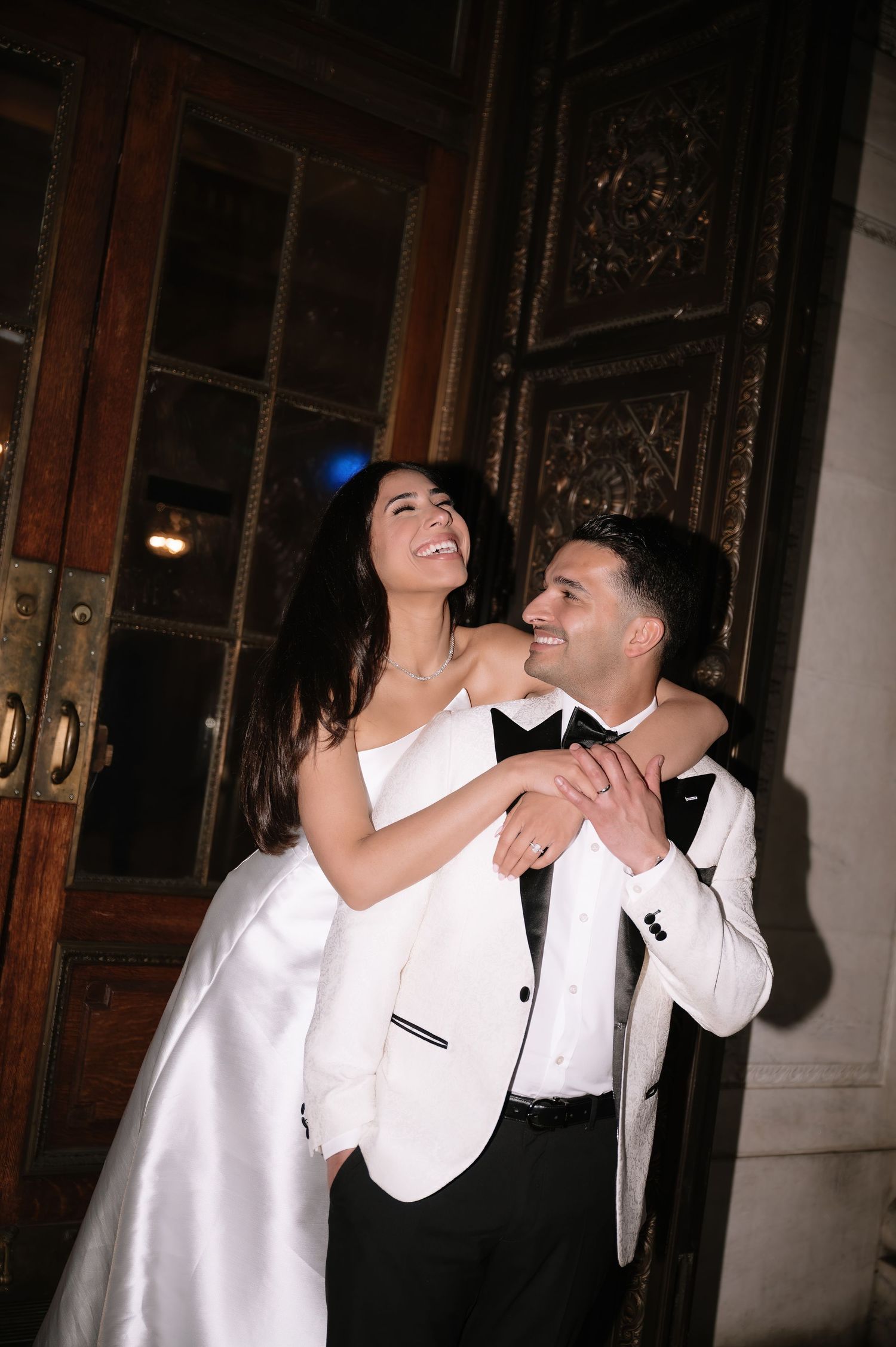 A joyful wedding couple laughs together while embracing in formal white attire near ornate wooden doors.