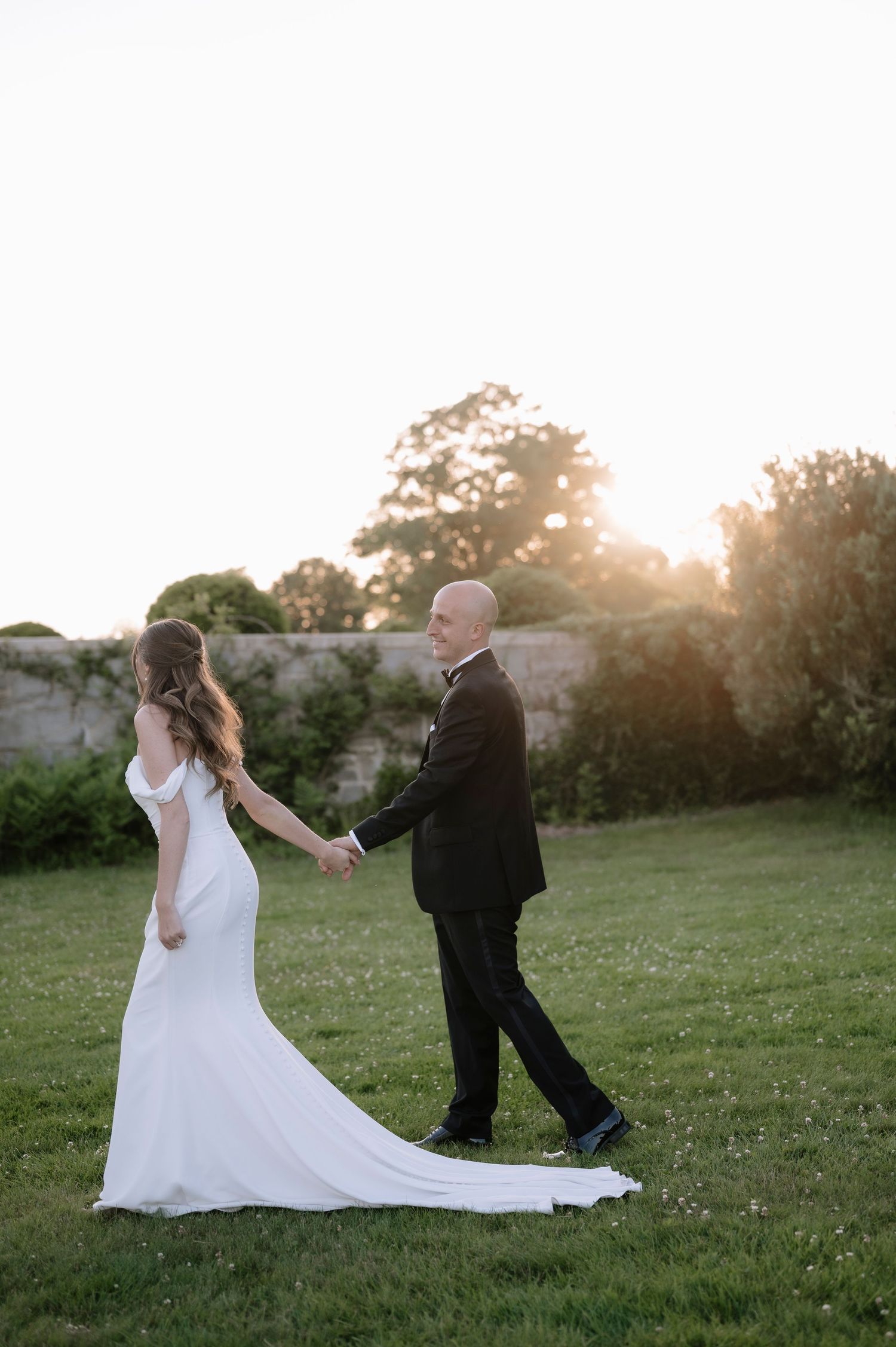 Newlywed couple walking on grass during sunset with bride in flowing white dress and groom in black suit.