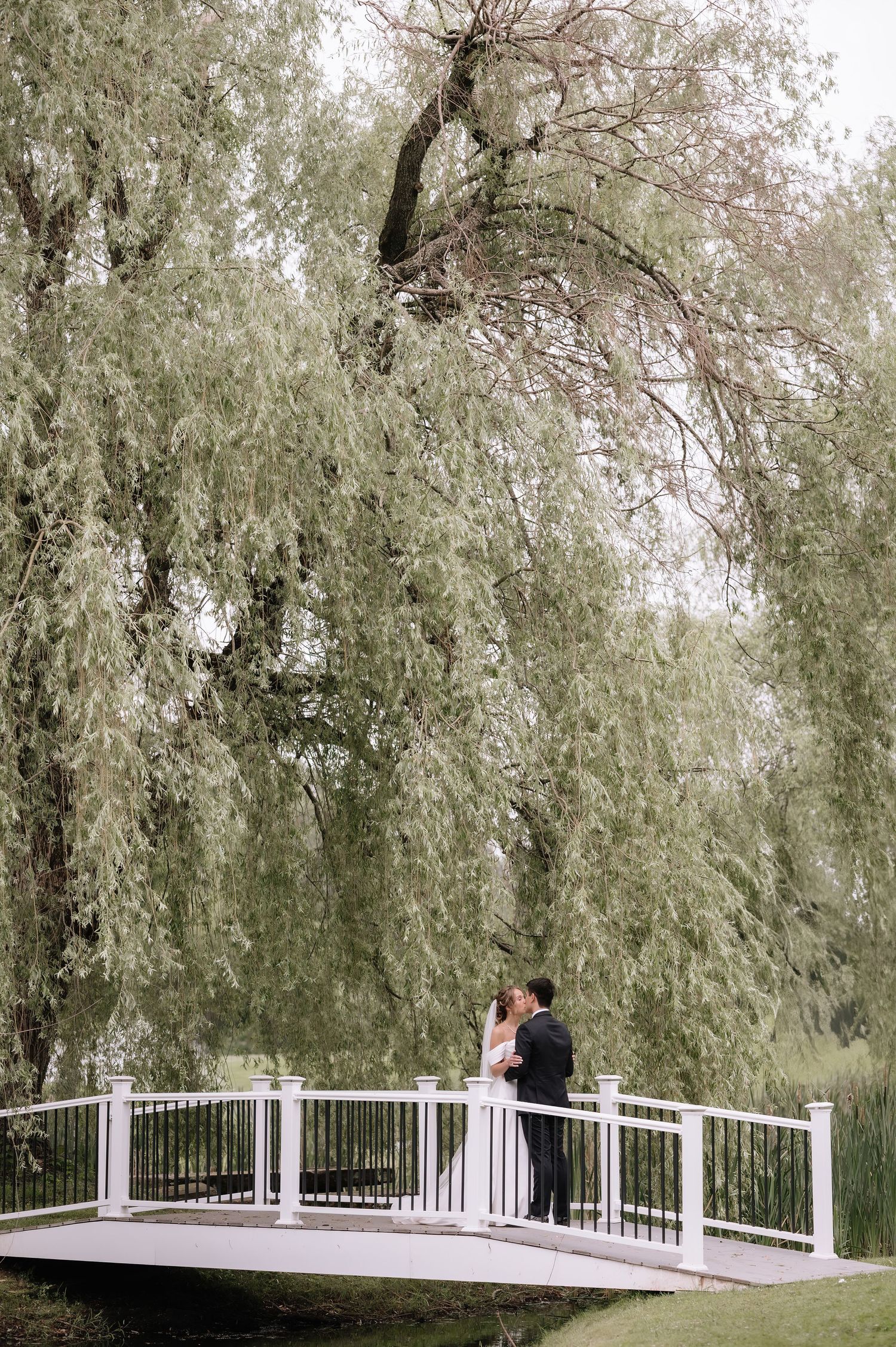 Romantic couple shares a tender moment on a white wooden bridge surrounded by graceful willow trees in a tranquil setting.