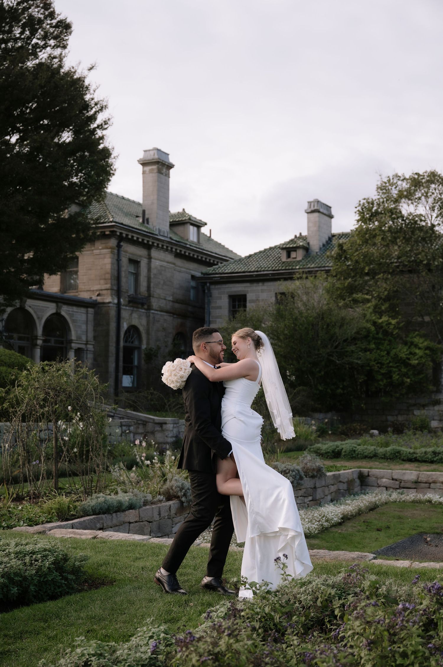 Elegant wedding photoshoot in a garden setting with stone mansion and landscaped grounds in background.