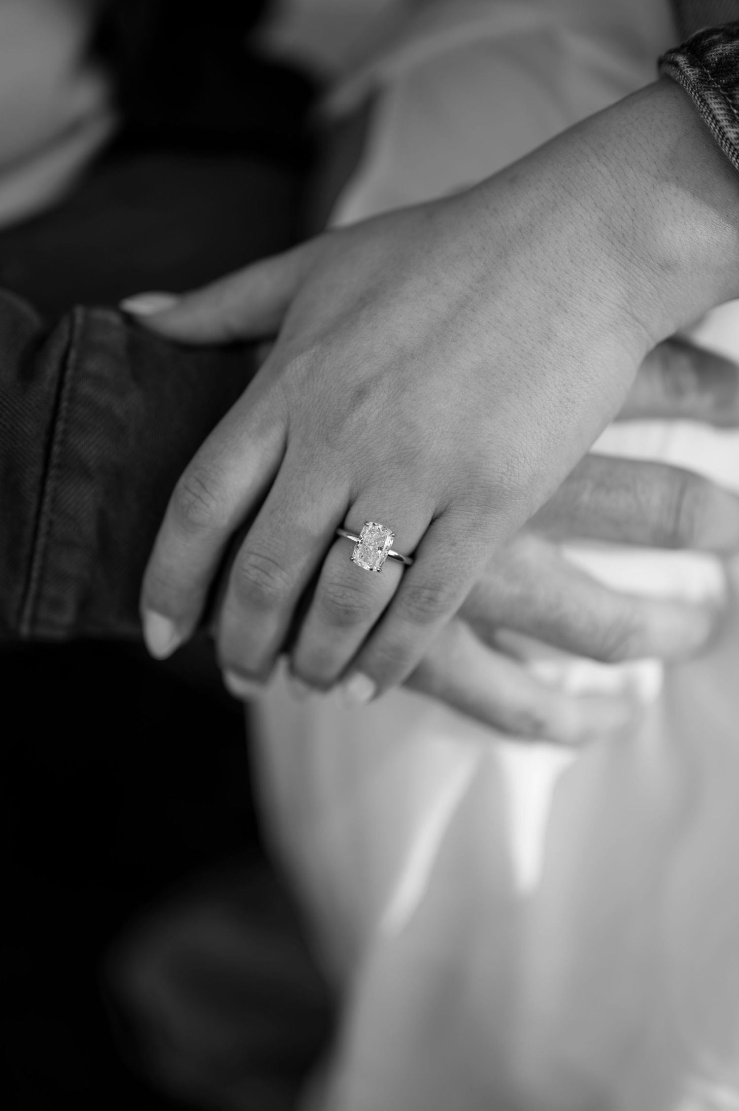Close-up black and white photo of a woman's hand wearing a stunning diamond engagement ring resting on a man's arm.