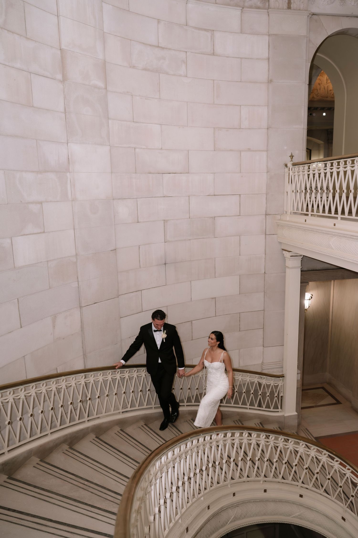Bride and groom holding hands on an elegant curved marble staircase in a grand wedding venue.