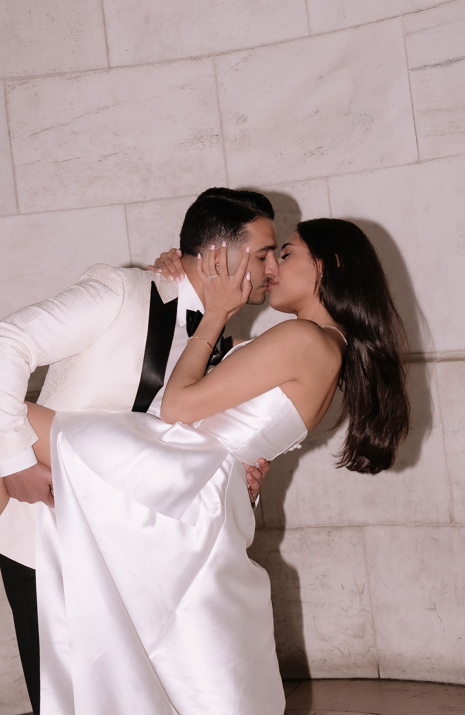 A romantic wedding couple shares a passionate kiss while the groom dips the bride in her elegant white dress against a wall.