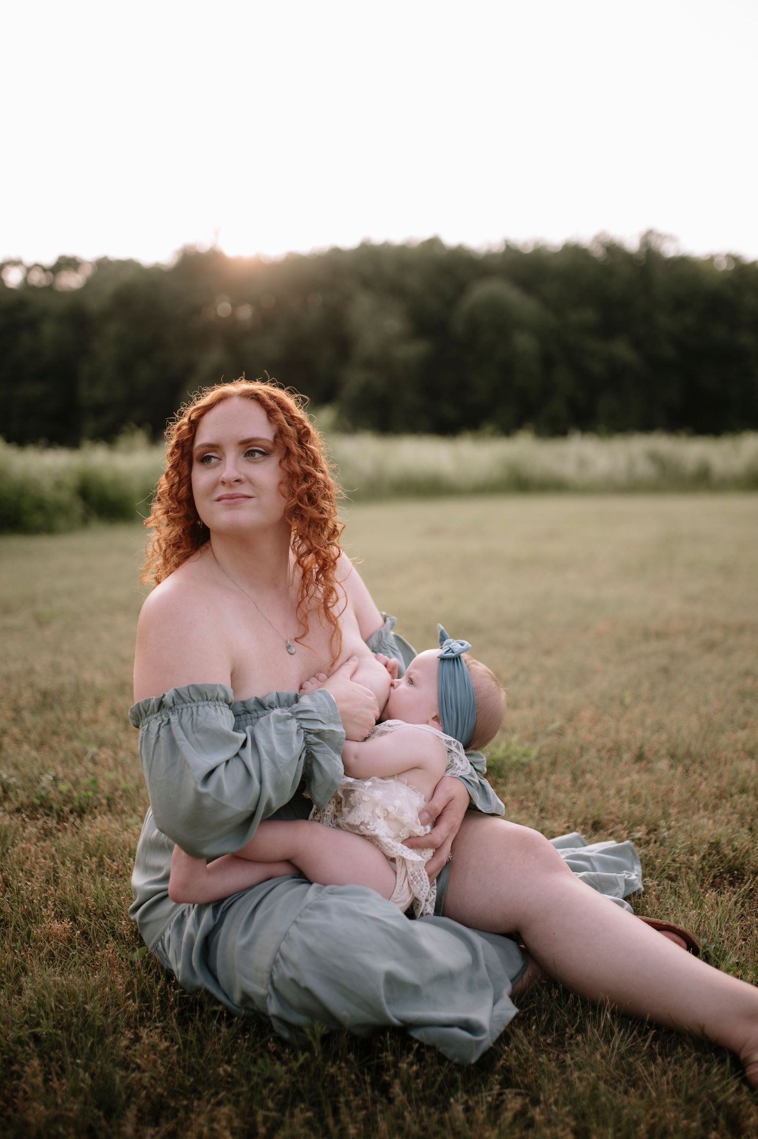 A mother nurses her baby outdoors in a sage green off-shoulder dress at sunset in a grassy field.