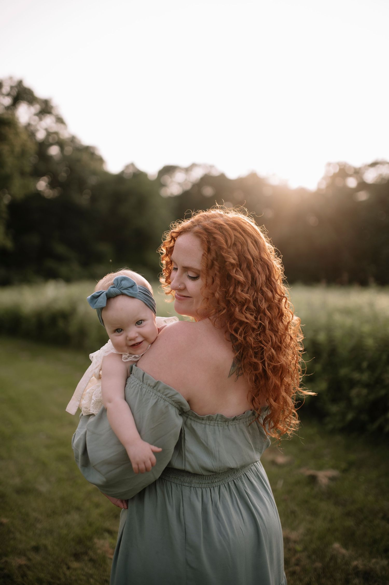 Beautiful sunset portrait of a woman in a sage green dress holding a smiling baby in a garden setting.