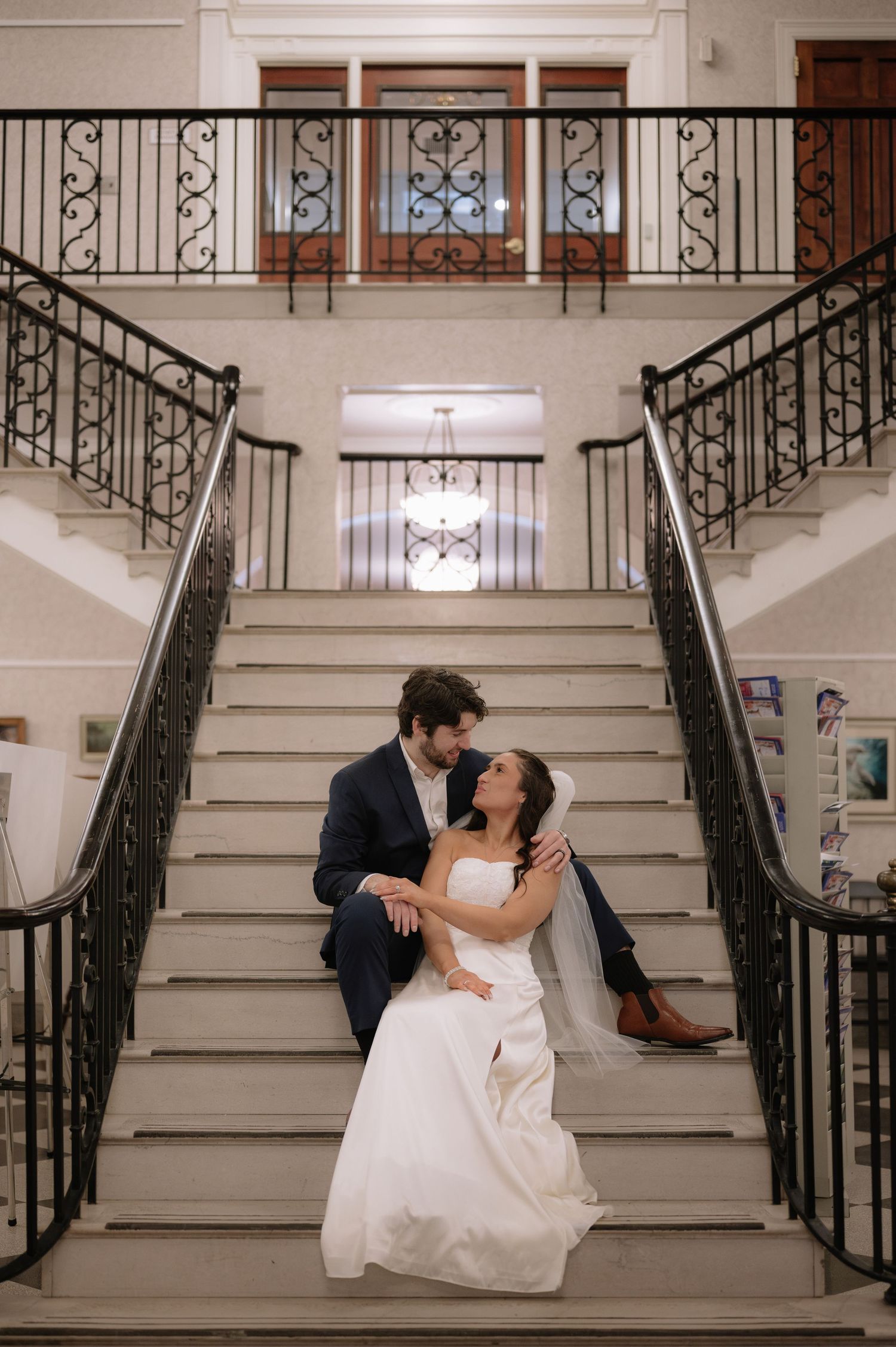 Couple in wedding attire share romantic moment on grand staircase with ornate iron railings.