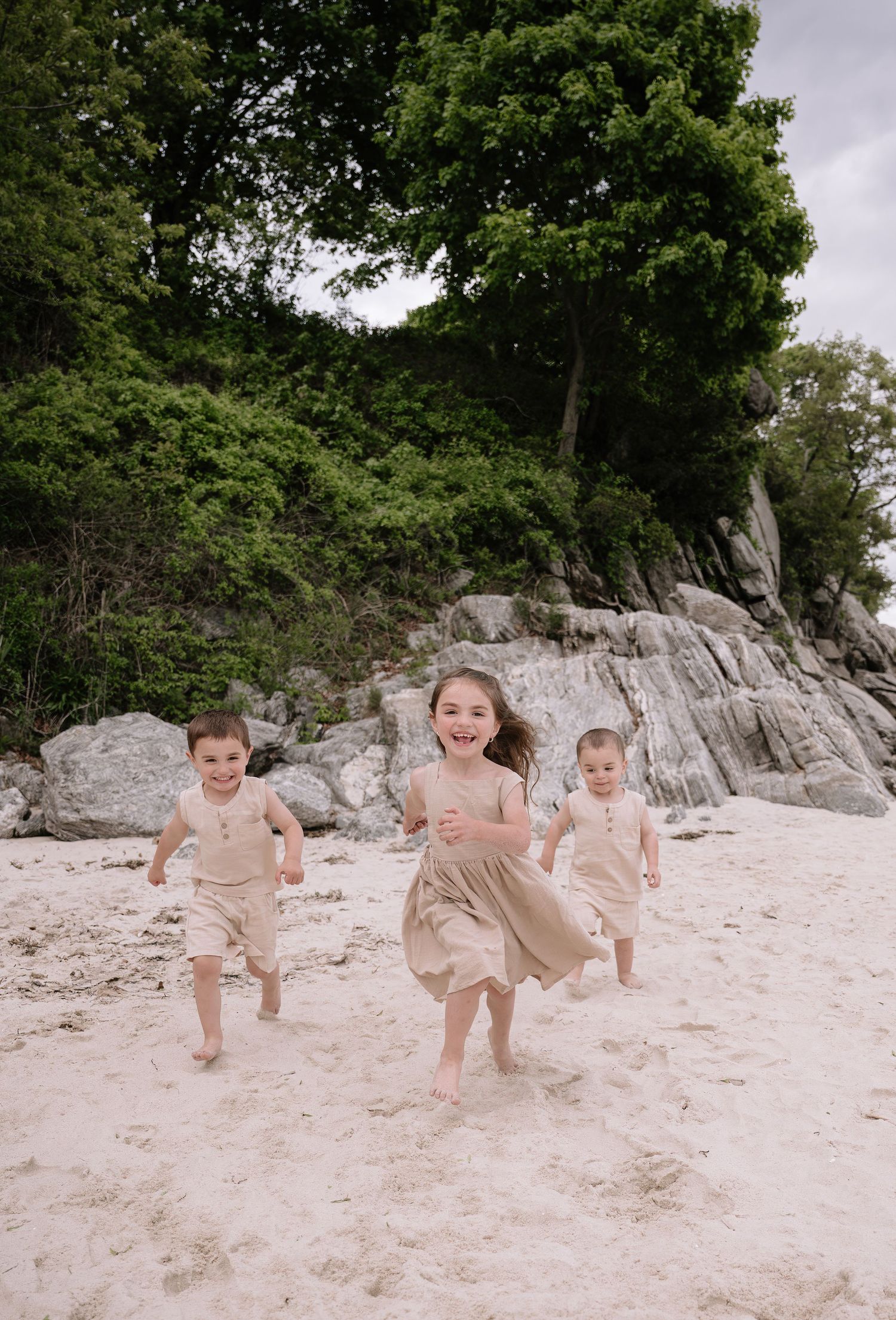 Three children in neutral clothing running and playing together on a sandy beach with rocky cliffs and trees in background.