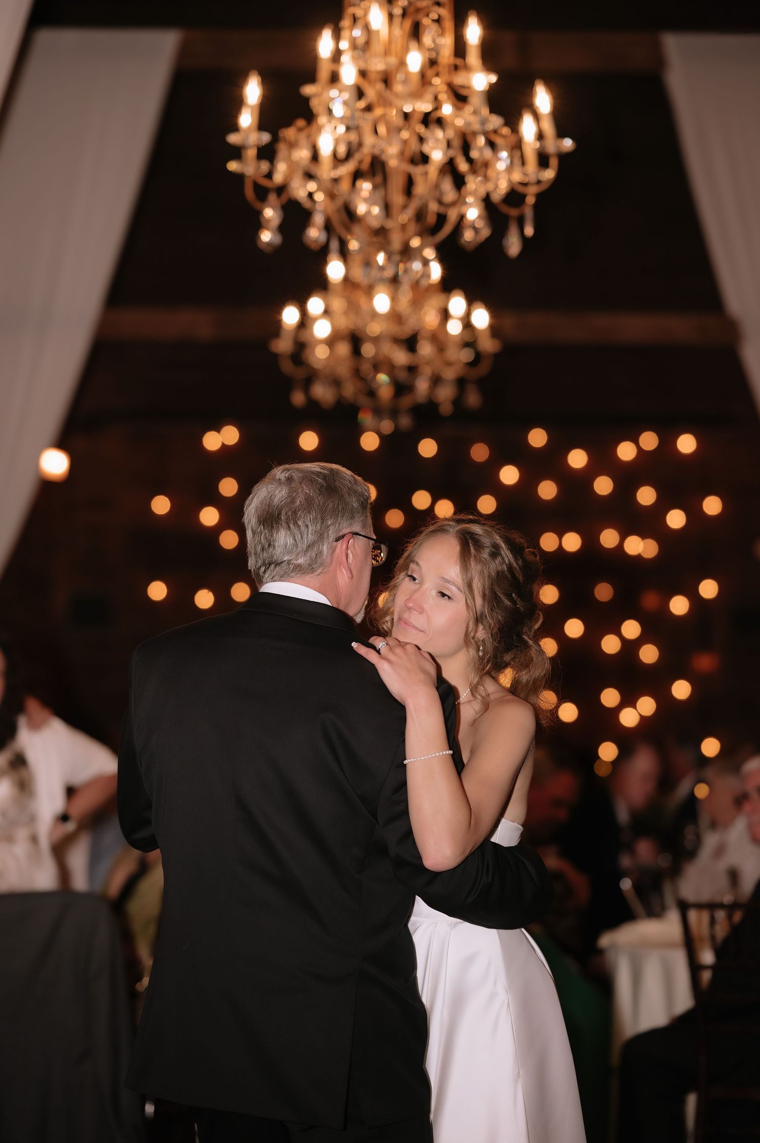Bride and father share an emotional first dance under a glowing chandelier at an elegant wedding reception.