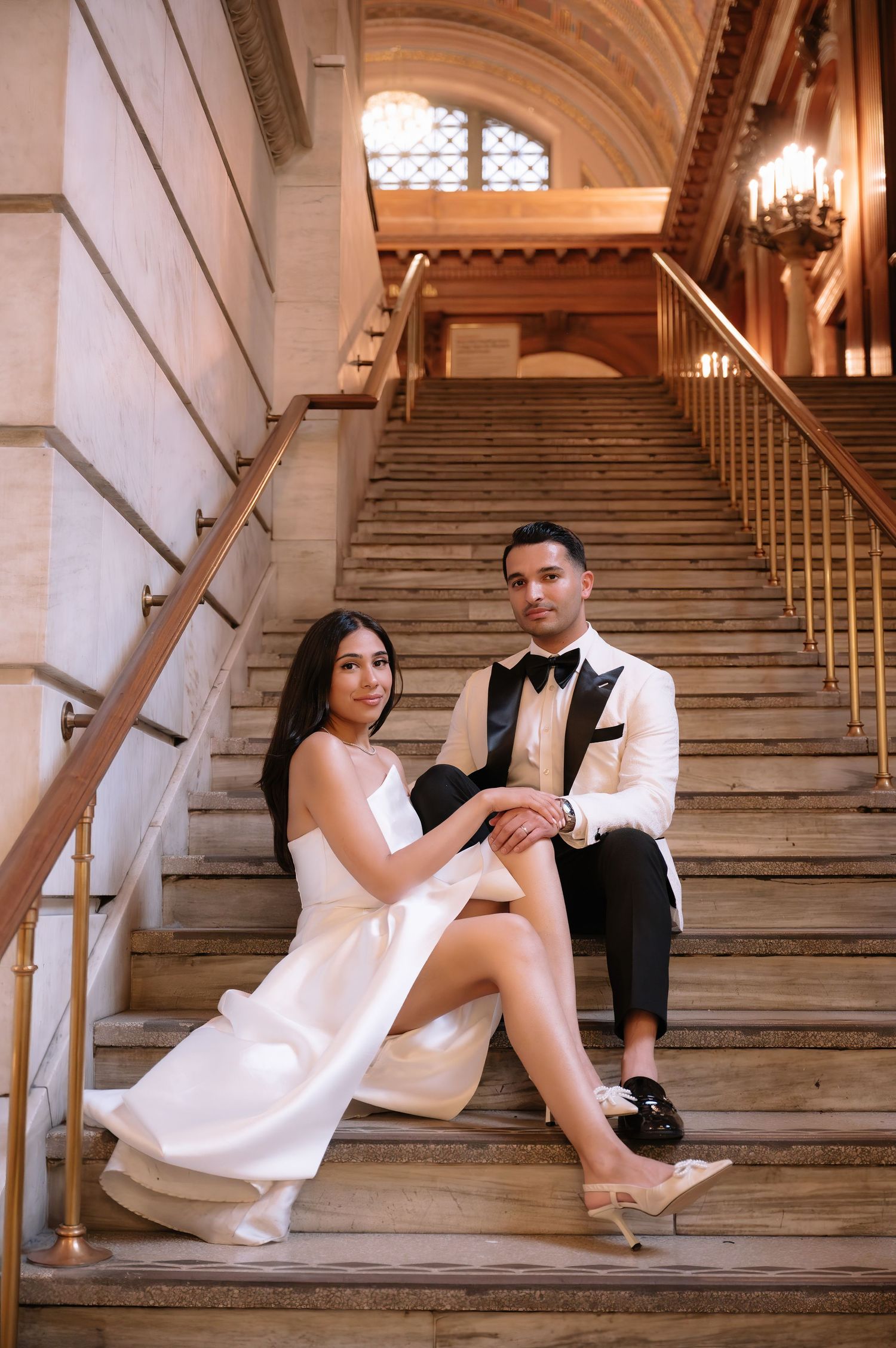 Elegant couple in formal attire pose on grand marble staircase with ornate architecture and chandelier in background.