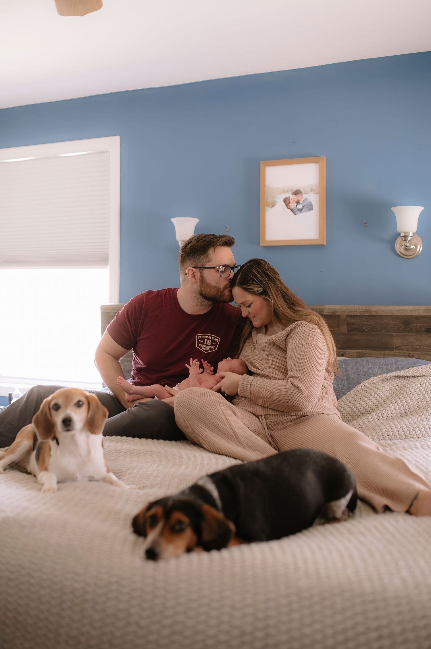 A couple relaxes on a bed with two dogs while sharing an intimate moment in a blue-walled bedroom.