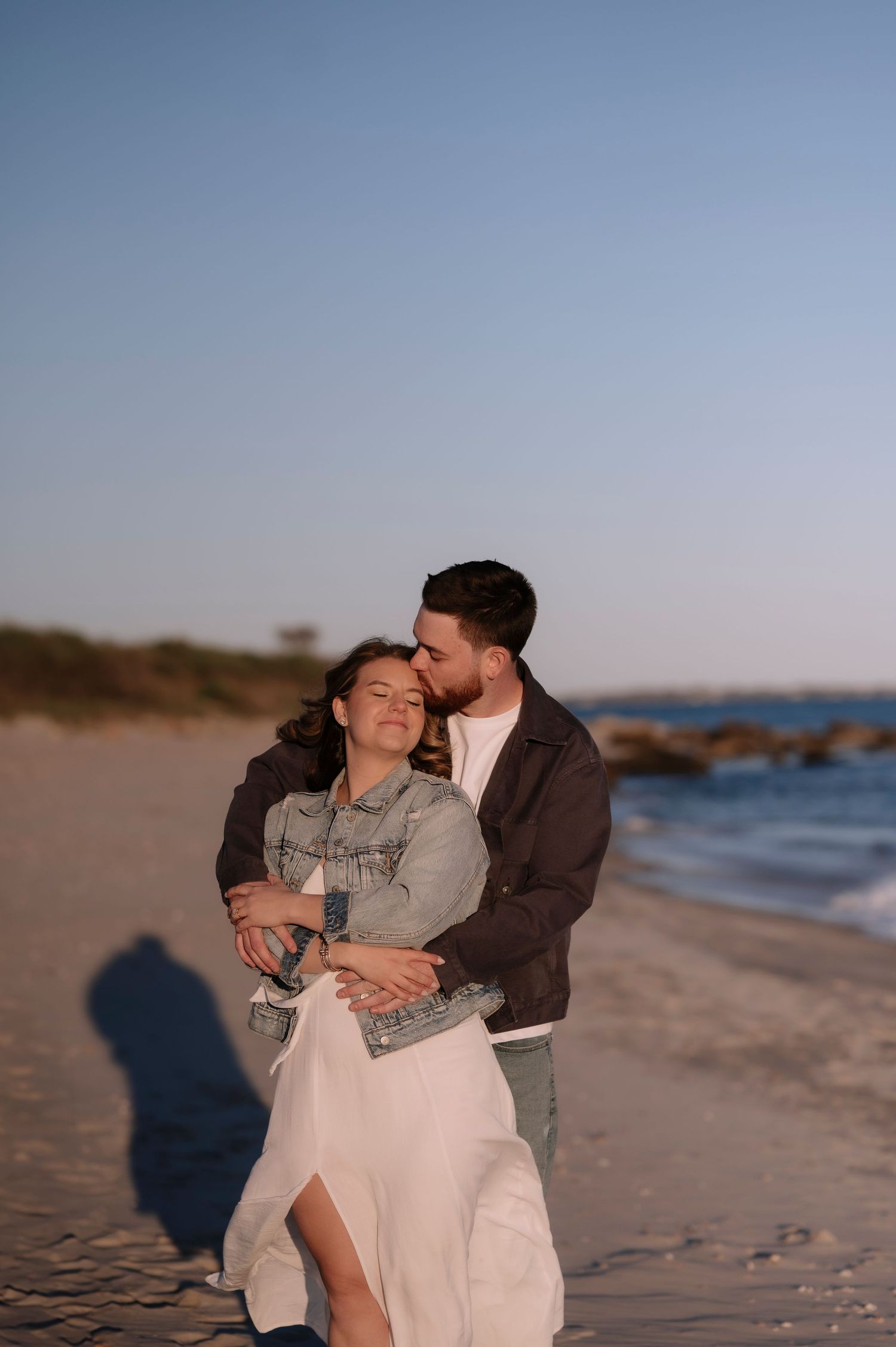 A couple embraces on a sandy beach at sunset, the man kissing the woman's temple as waves roll in behind them.
