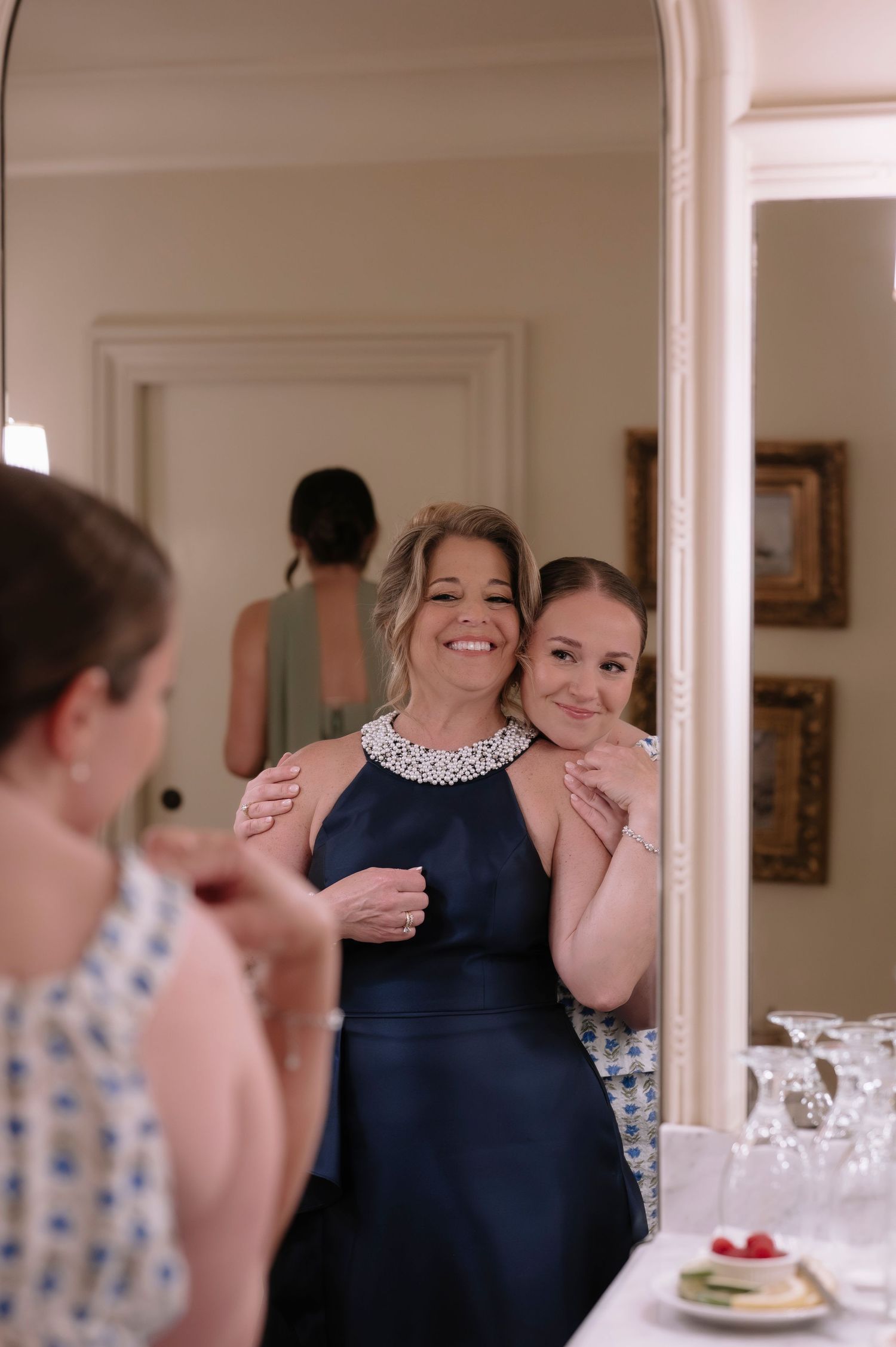 Two women smile together in a mirror reflection, one in a navy halter dress with beaded neckline, at a wedding preparation.