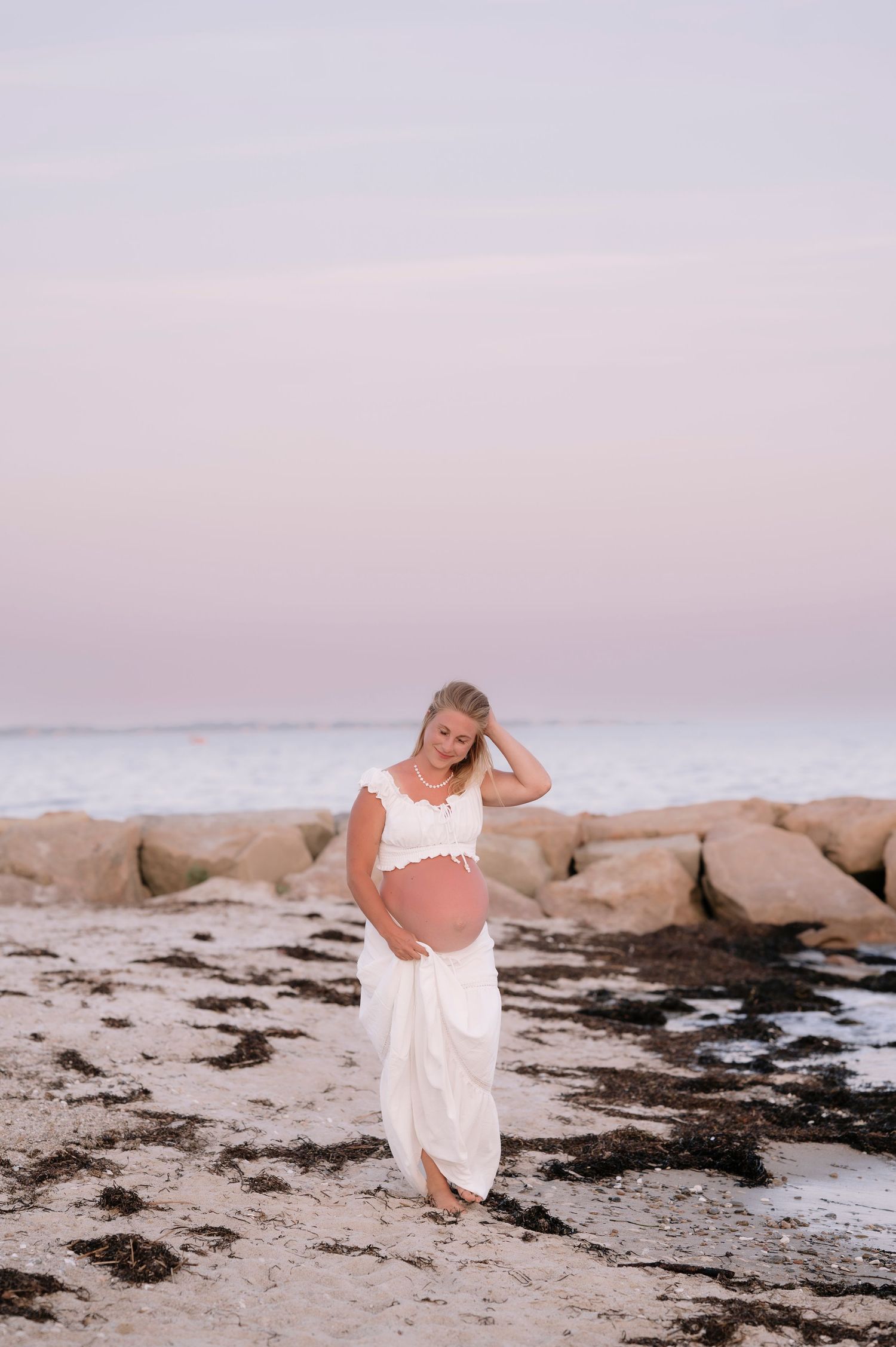 Pregnant woman in white two-piece outfit standing on rocky beach at sunset, holding her baby bump.