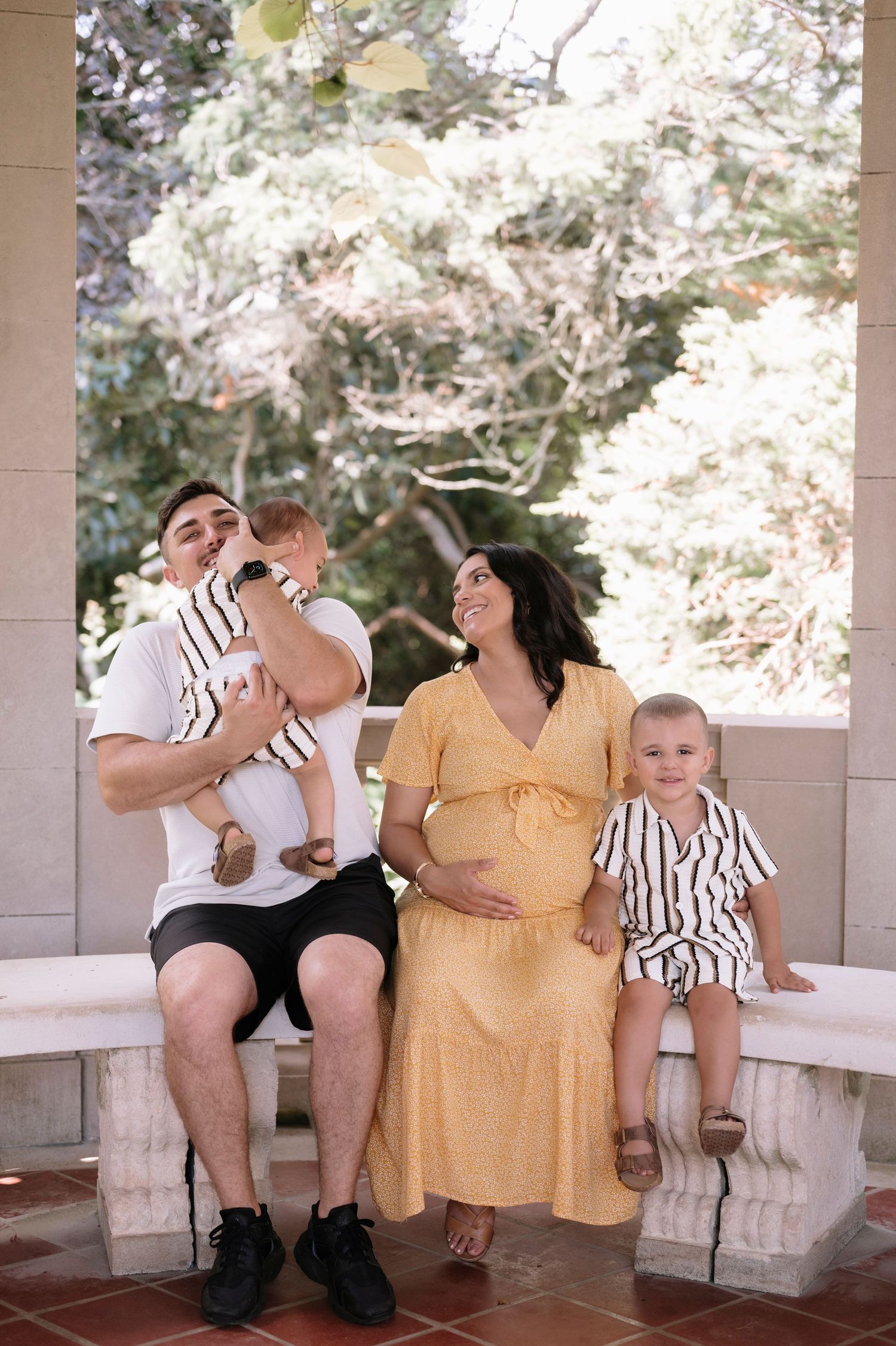 Pregnant woman in yellow dress sits with partner holding baby and toddler on stone bench outdoors.