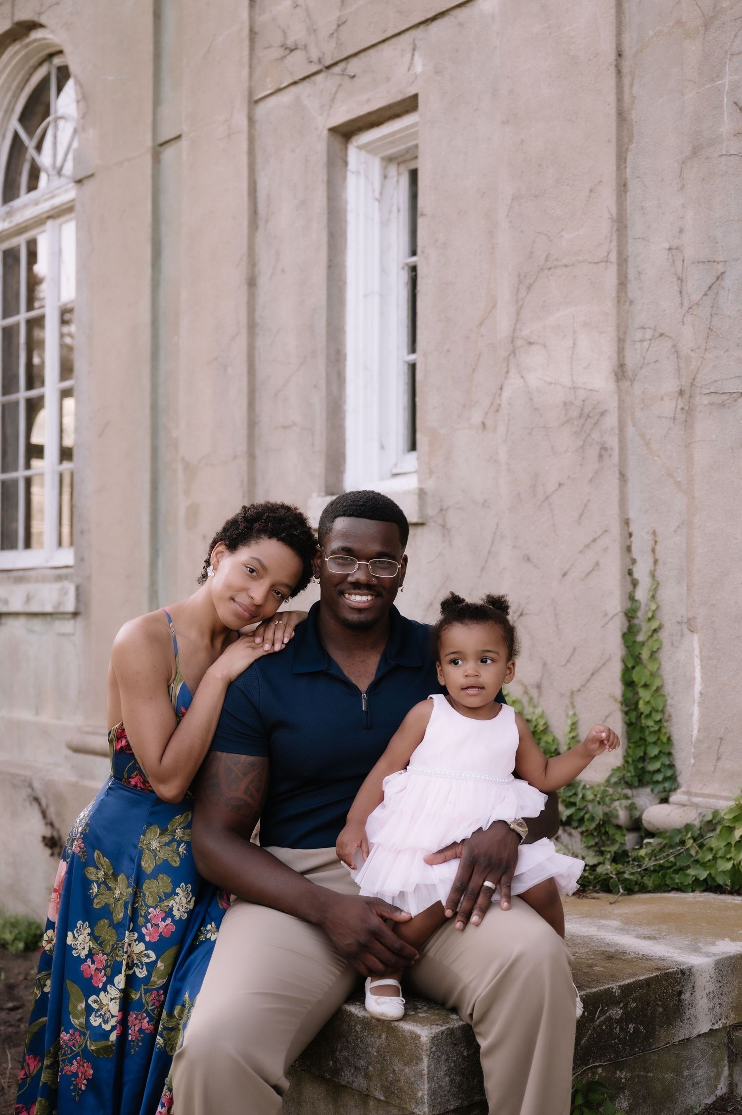 A happy Black family of three poses outdoors, parents smiling with their baby girl in a white dress.