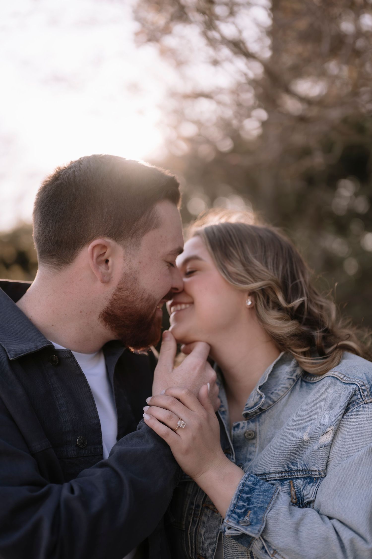 A couple shares a romantic moment outdoors, smiling and holding hands, with a diamond engagement ring visible in golden sunlight.