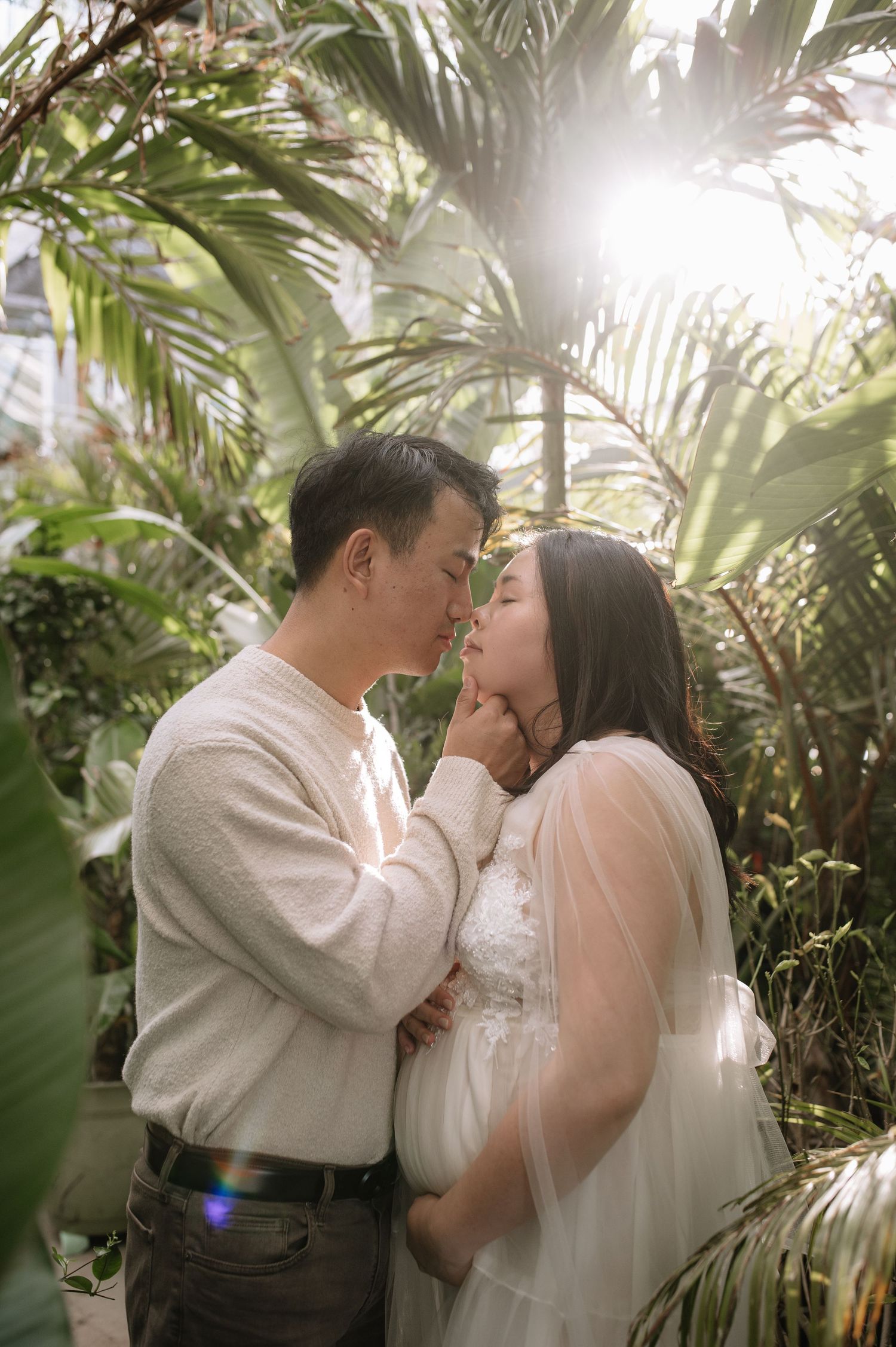 A tender maternity moment captured in a greenhouse with sunlight streaming through palm fronds.