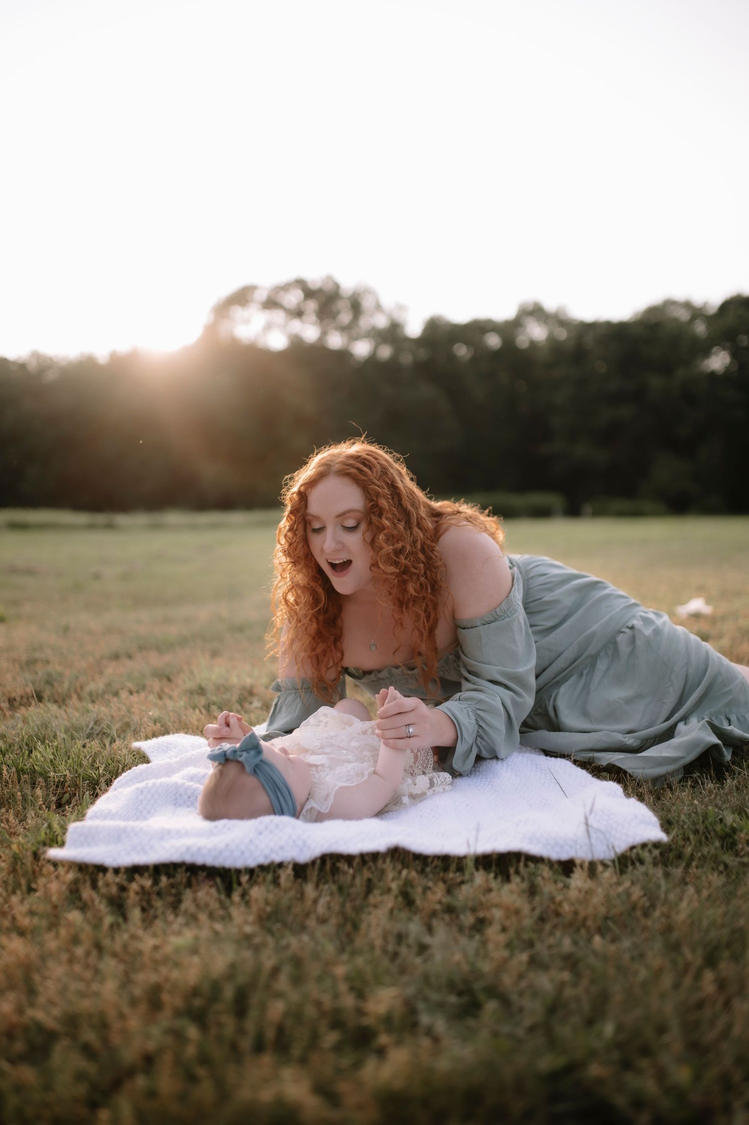 A mother and infant share a tender moment on a blanket in a sunset-lit grassy field.