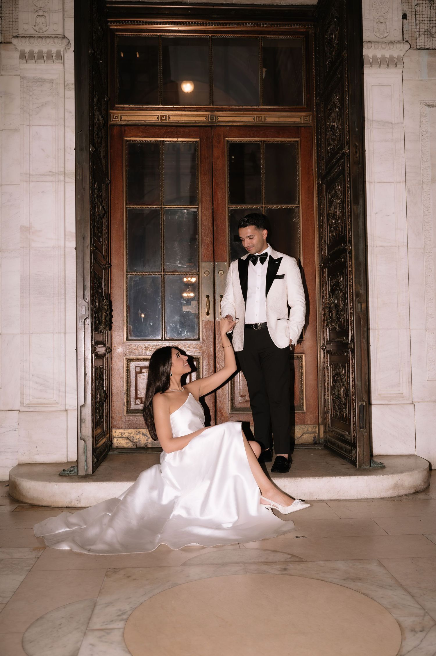 A romantic wedding photo of a couple in formal attire posed elegantly in front of ornate vintage wooden doors.