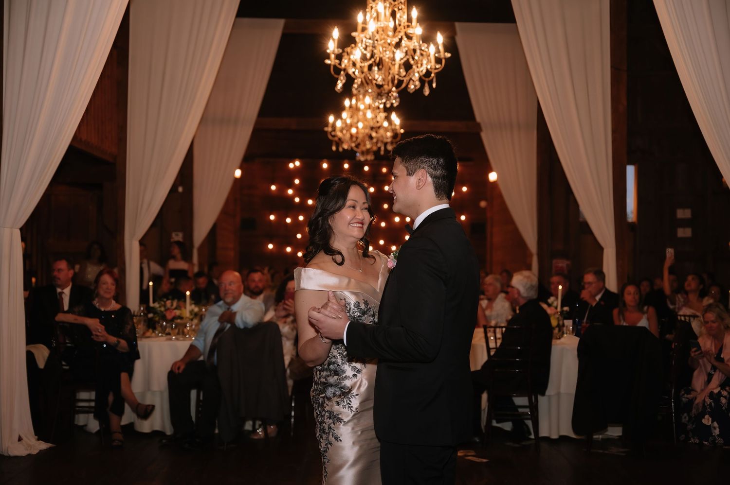 Couple shares first dance under chandelier at elegant wedding reception with guests watching in candlelit venue.