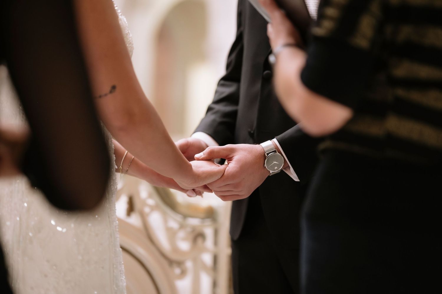 A couple holds hands tenderly at a wedding ceremony, the groom wearing a silver watch and dark suit.