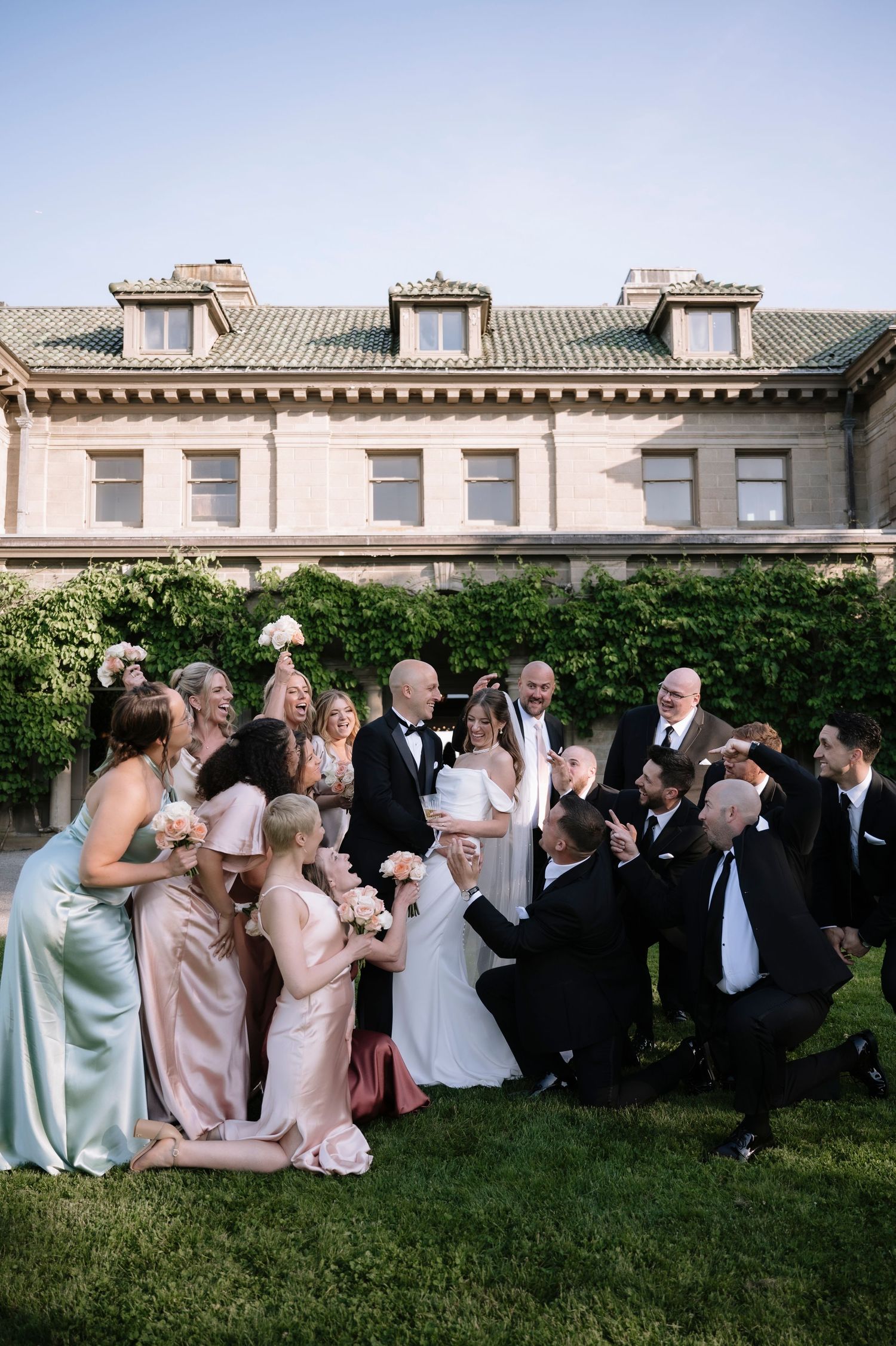 Wedding party gathers on the lawn of an elegant stone mansion with dormers during an outdoor celebration.
