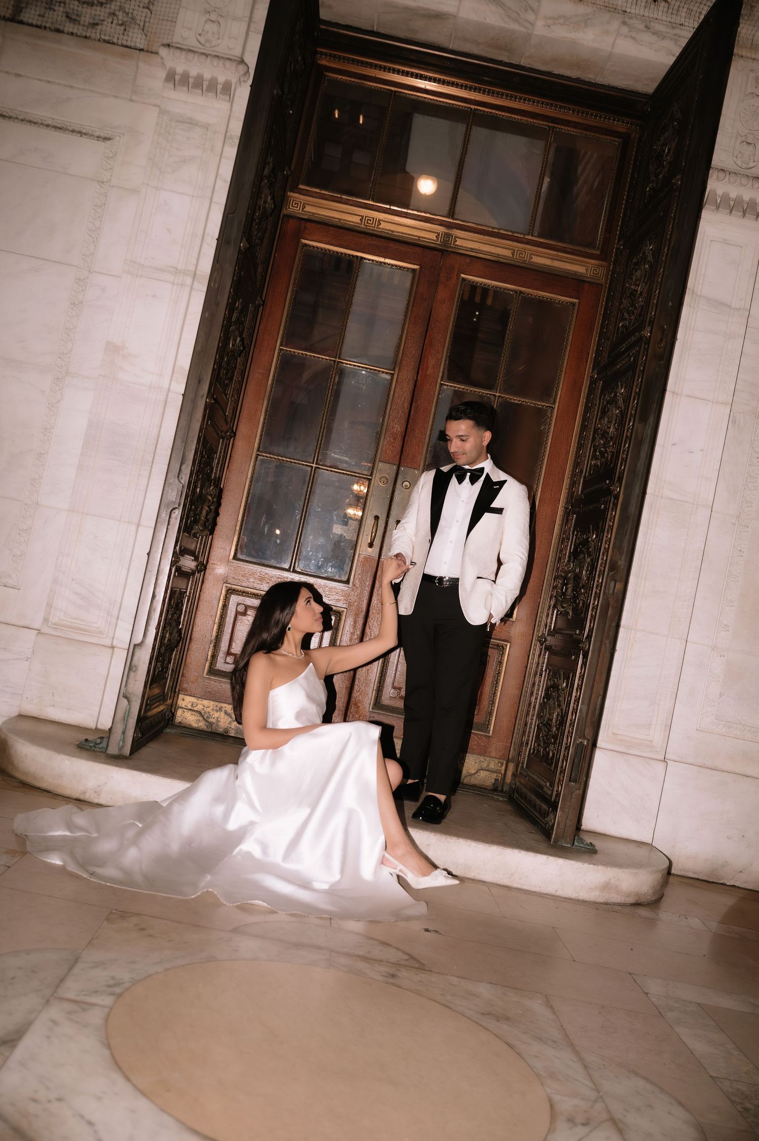 A bride in a white gown and person in white tuxedo pose elegantly by ornate wooden doors in a classic building interior.