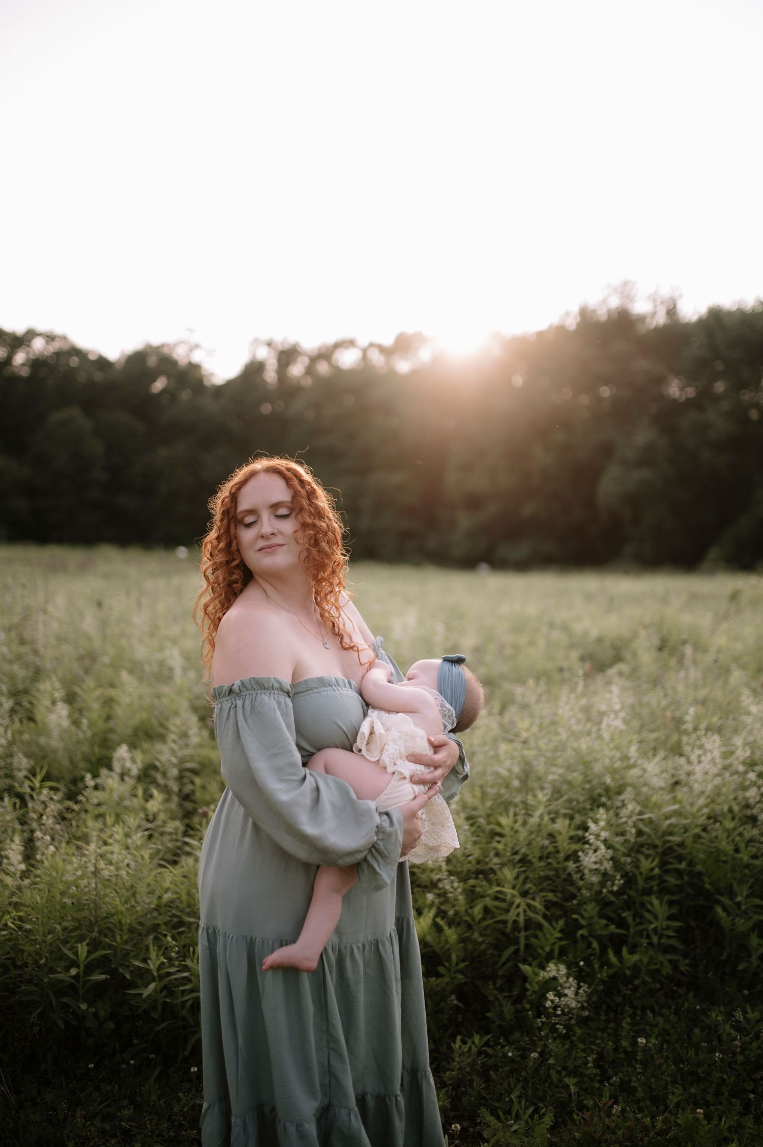 Mother in flowing sage green dress holds infant in golden sunset light while standing in wildflower meadow.