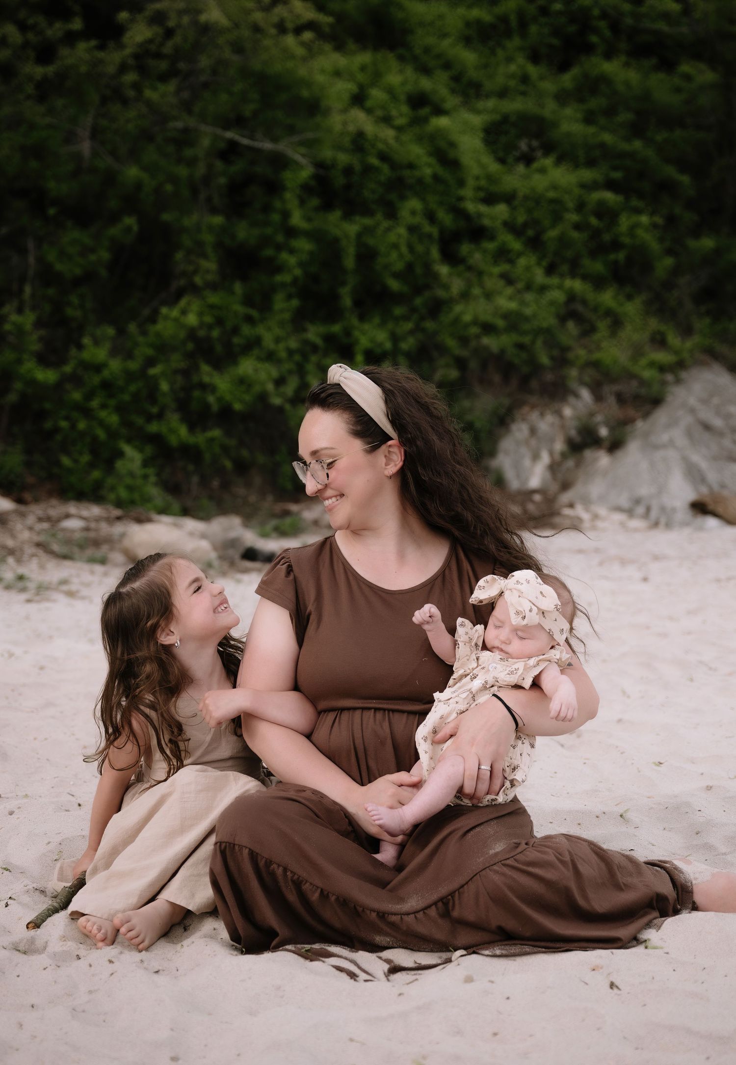 A family shares a tender moment together while sitting on a sandy beach against a backdrop of lush greenery.
