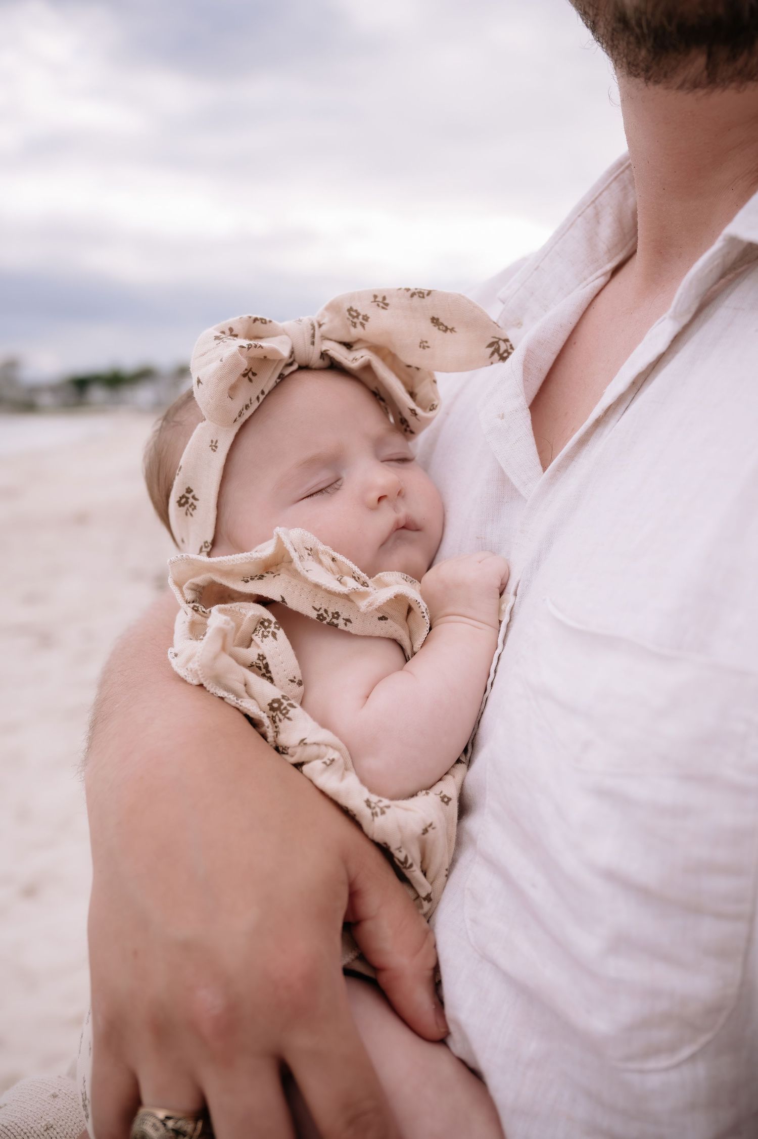 A sleeping infant wearing a beige headband rests peacefully against someone's chest at the beach.