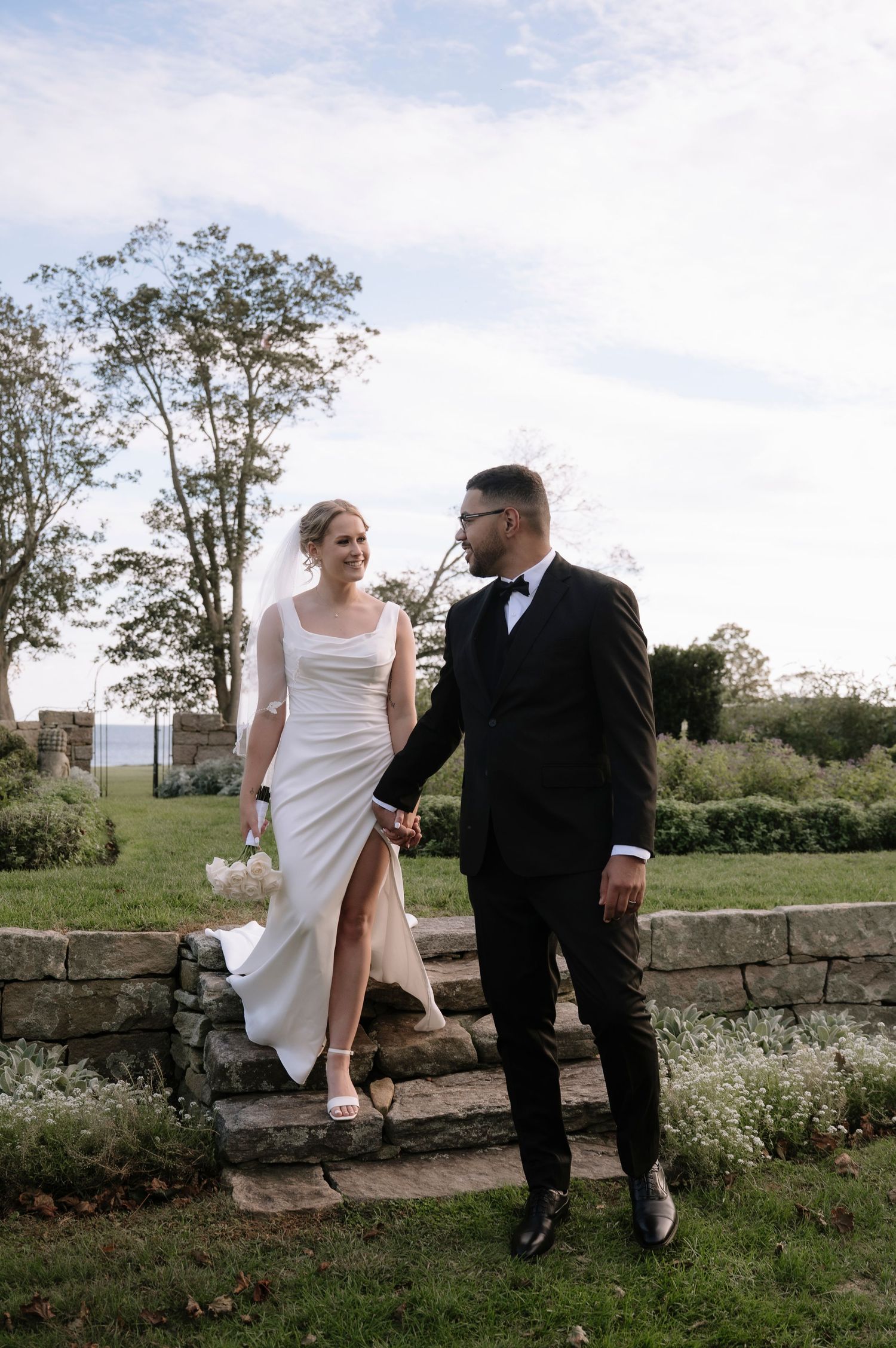 A couple in wedding attire walks together on stone steps in a scenic outdoor garden setting.