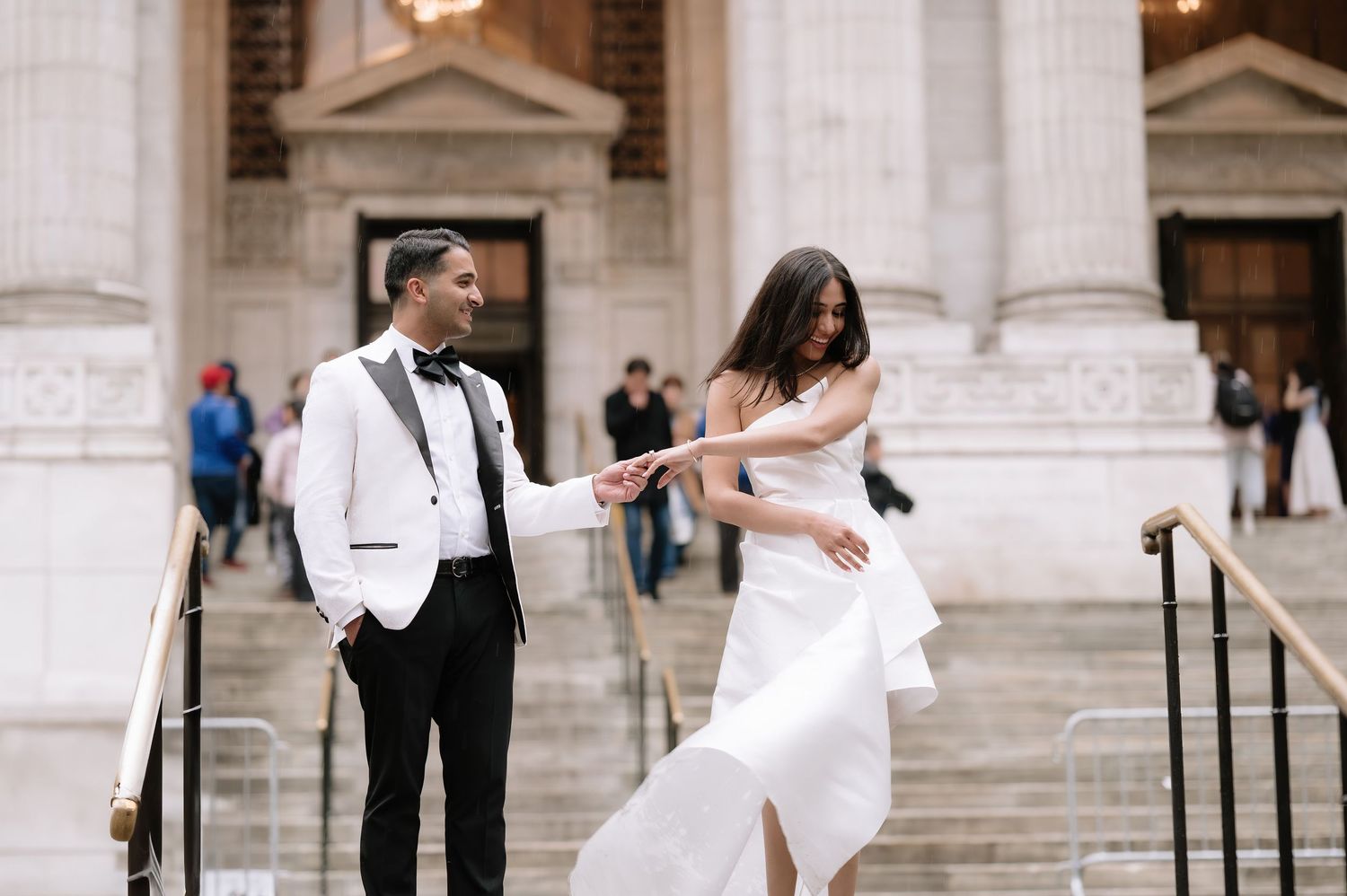 A couple in wedding attire holds hands on grand stone steps, groom in white tuxedo jacket, bride in elegant white gown.
