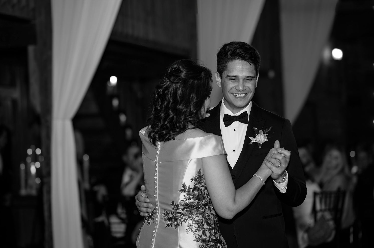 Black and white photo of a couple sharing their first dance at a wedding reception, groom smiling in a tuxedo.