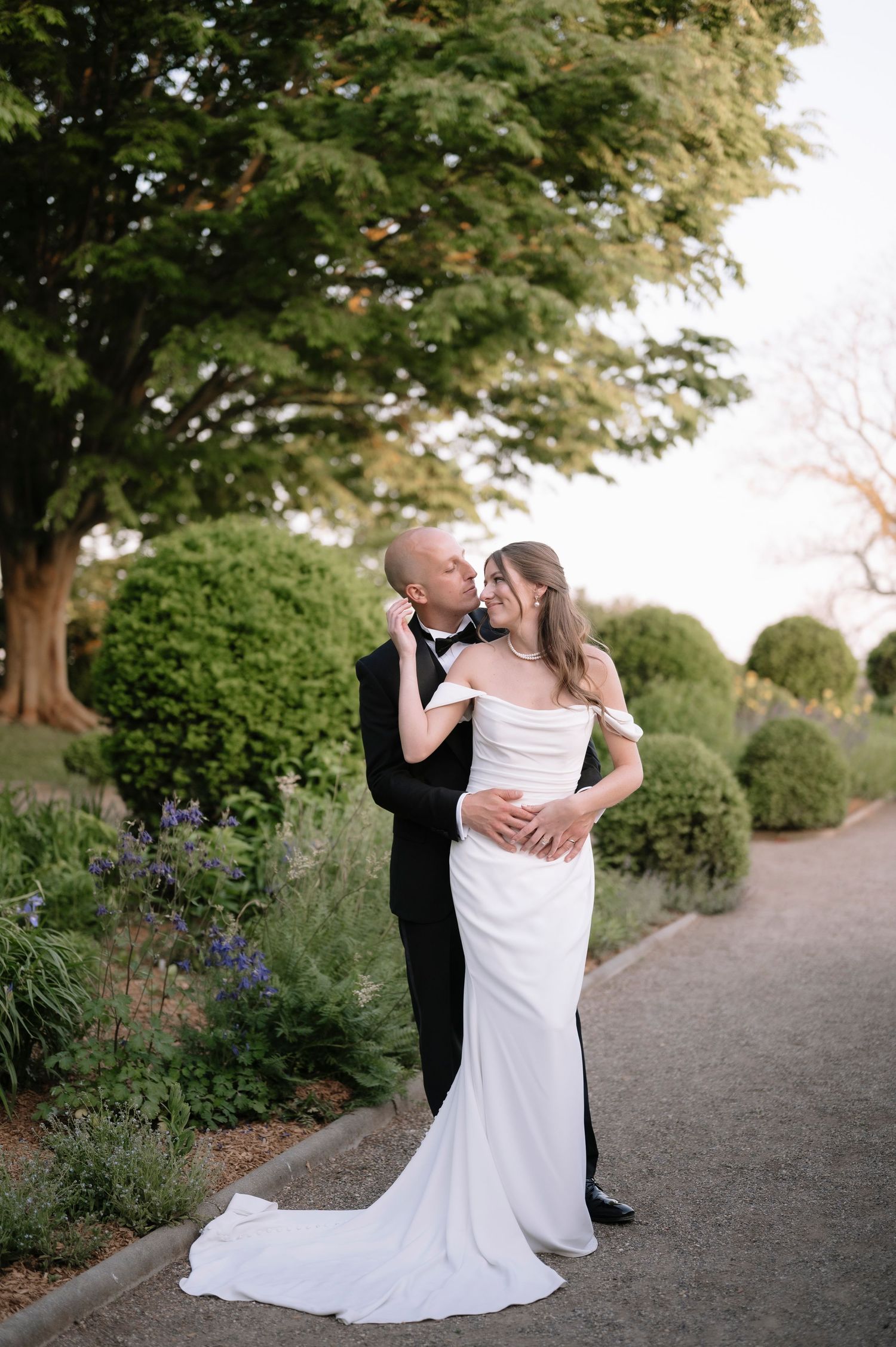 Newlyweds share a romantic embrace on a garden path lined with lush greenery at sunset.