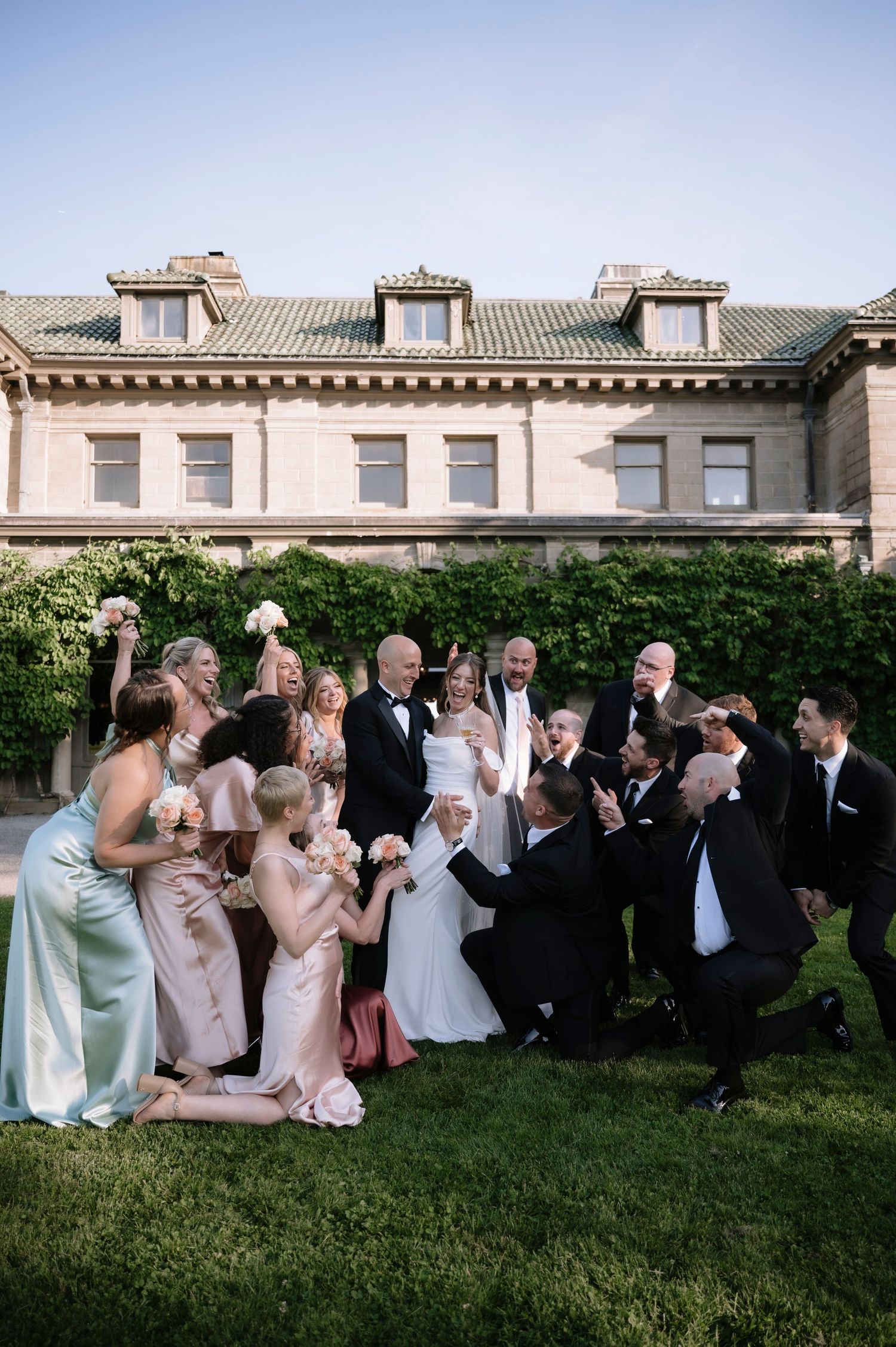 Wedding party gathered on mansion lawn striking playful poses in formal attire against historic stone building backdrop.