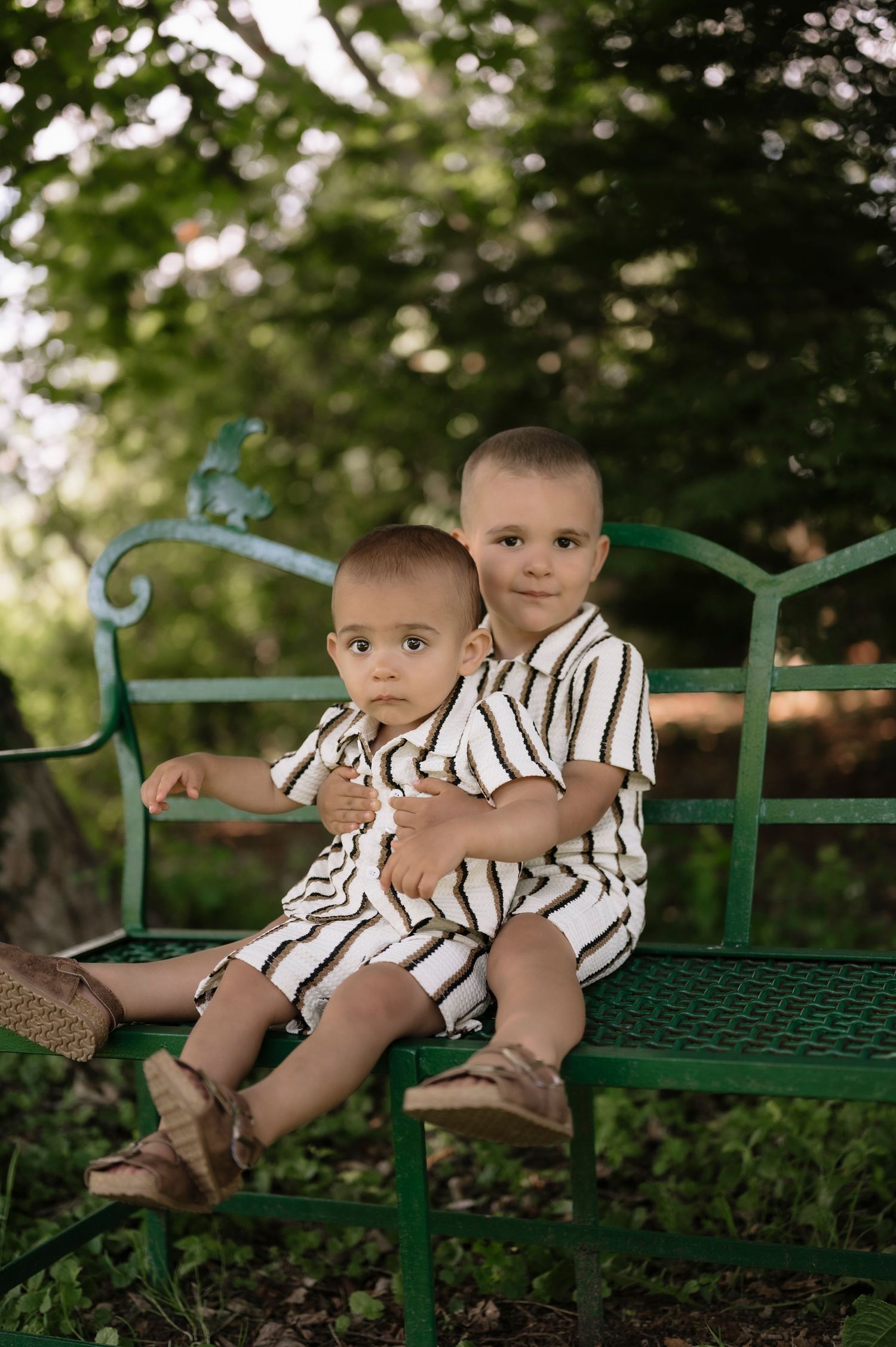 Two young brothers in matching striped outfits sit together on a green metal bench outdoors surrounded by lush trees.