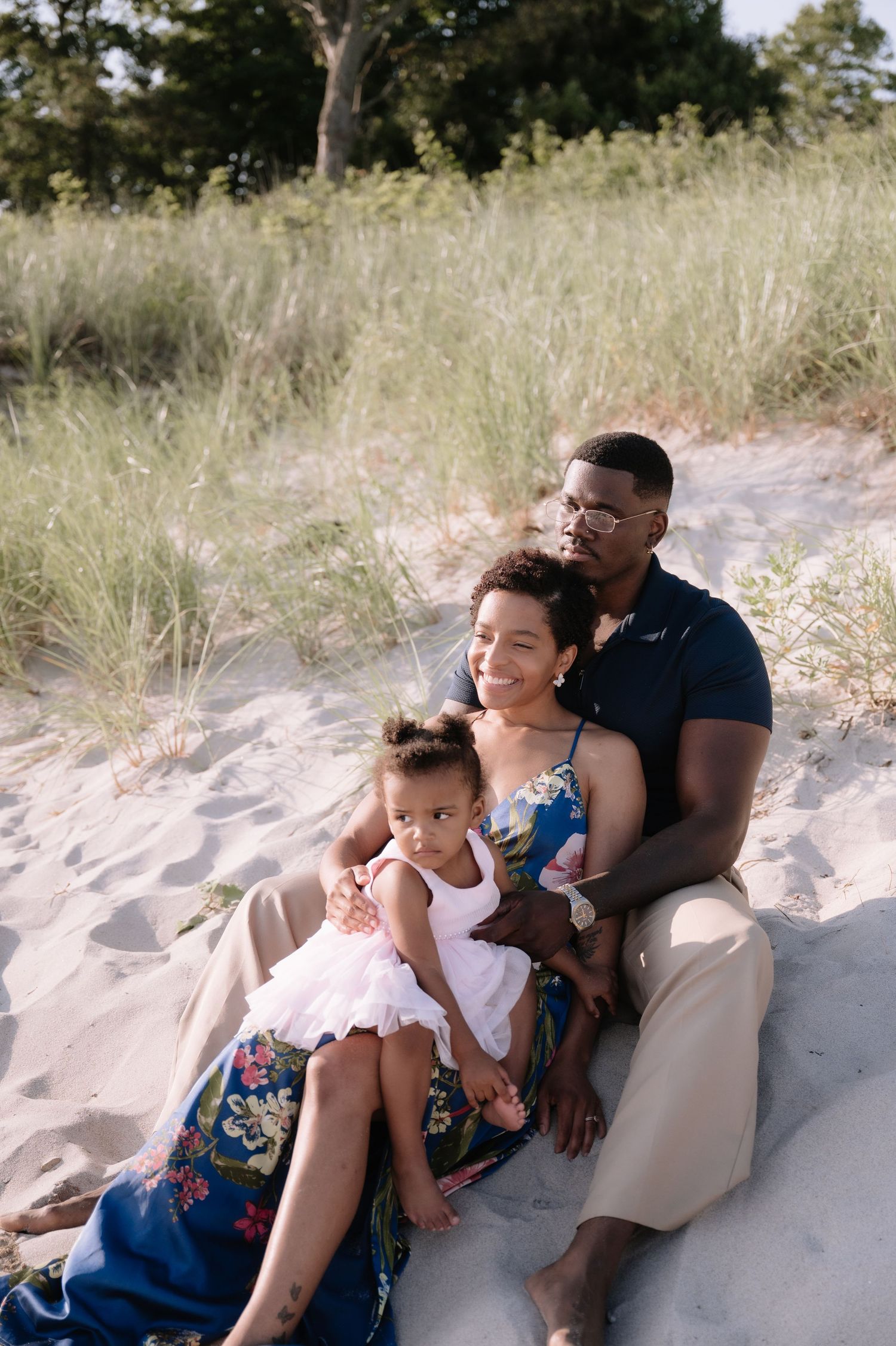 A family of three sits together on sandy beach dunes, with a man, woman, and young toddler girl enjoying a sunny day.