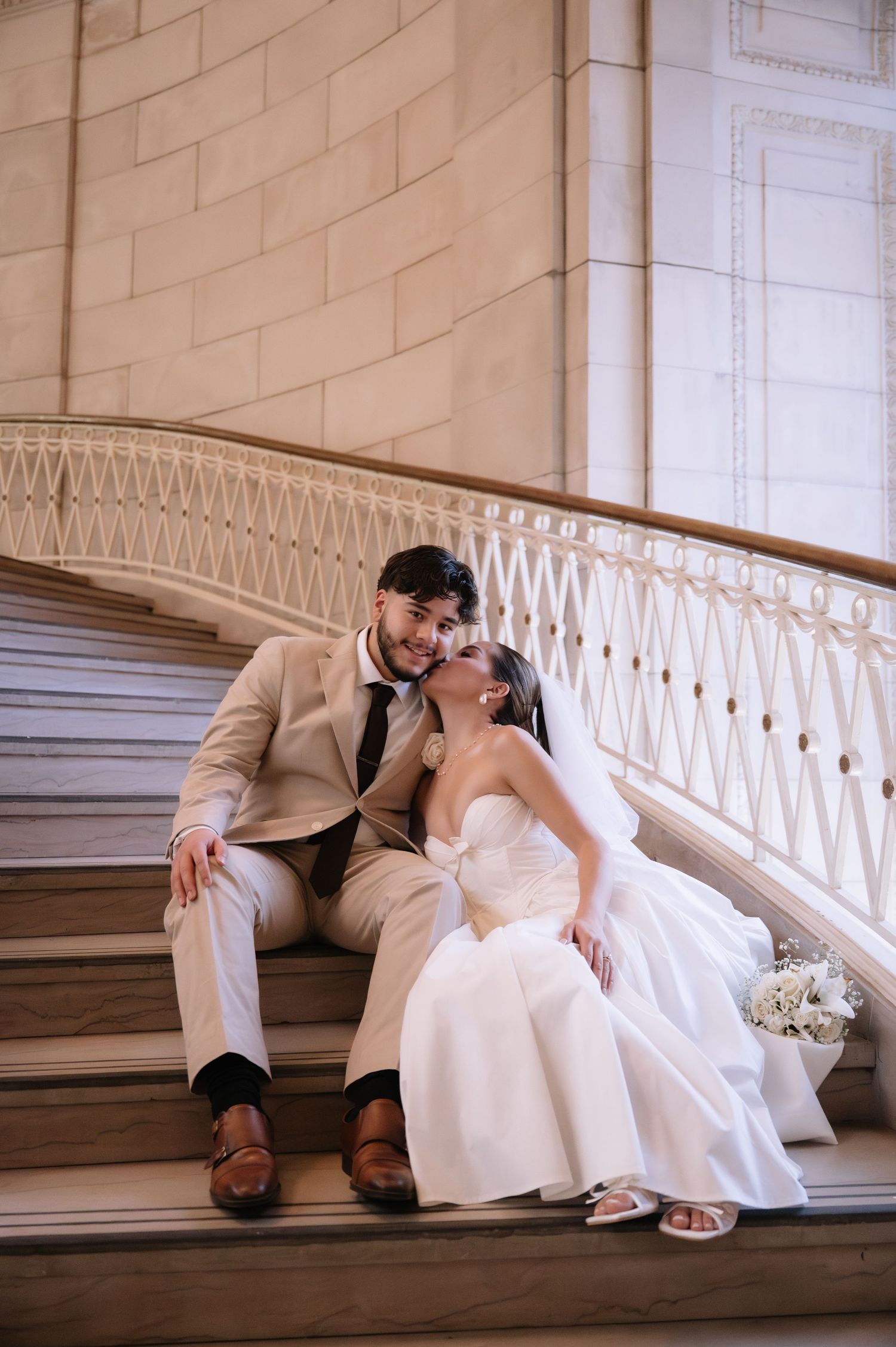Romantic couple in wedding attire lounging on grand marble stairs in an elegant architectural setting.