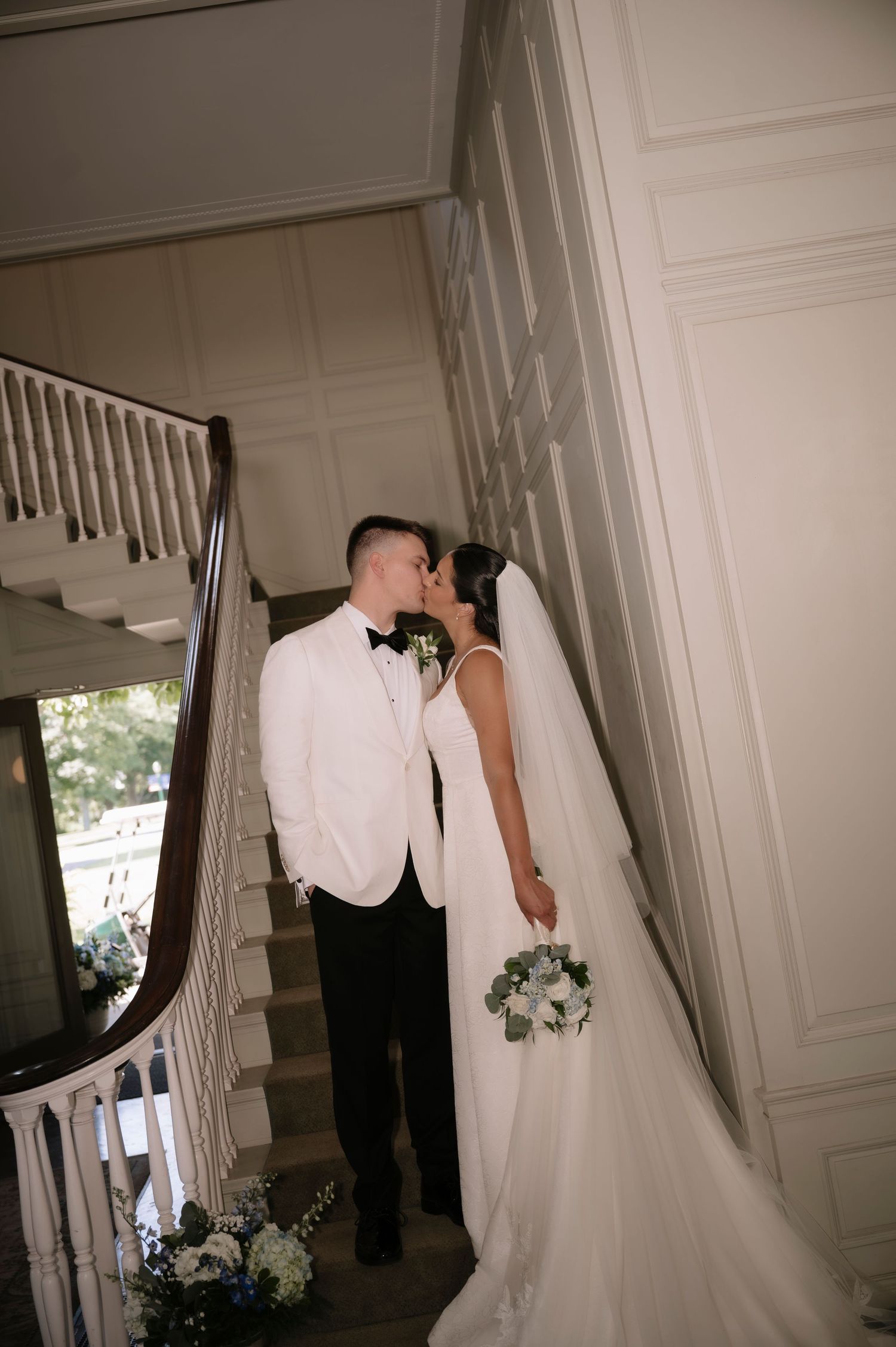 Bride and groom share a kiss on elegant staircase, bride holding white bouquet, groom in white tuxedo jacket.