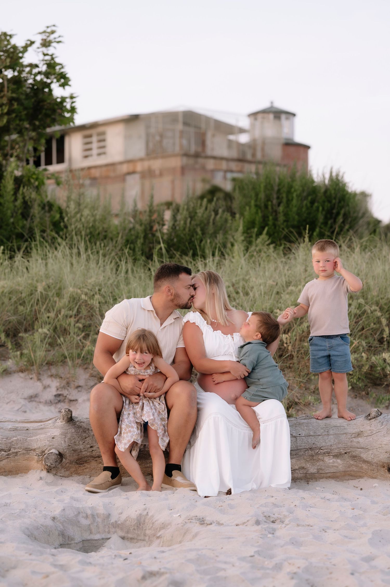 Family of five sharing a tender moment on sandy beach, parents kissing while children play around them.