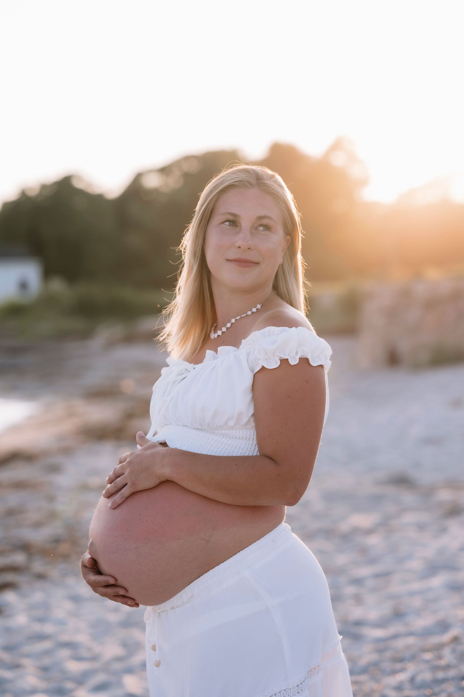 Pregnant woman in white dress cradling baby bump on beach at golden hour sunset.